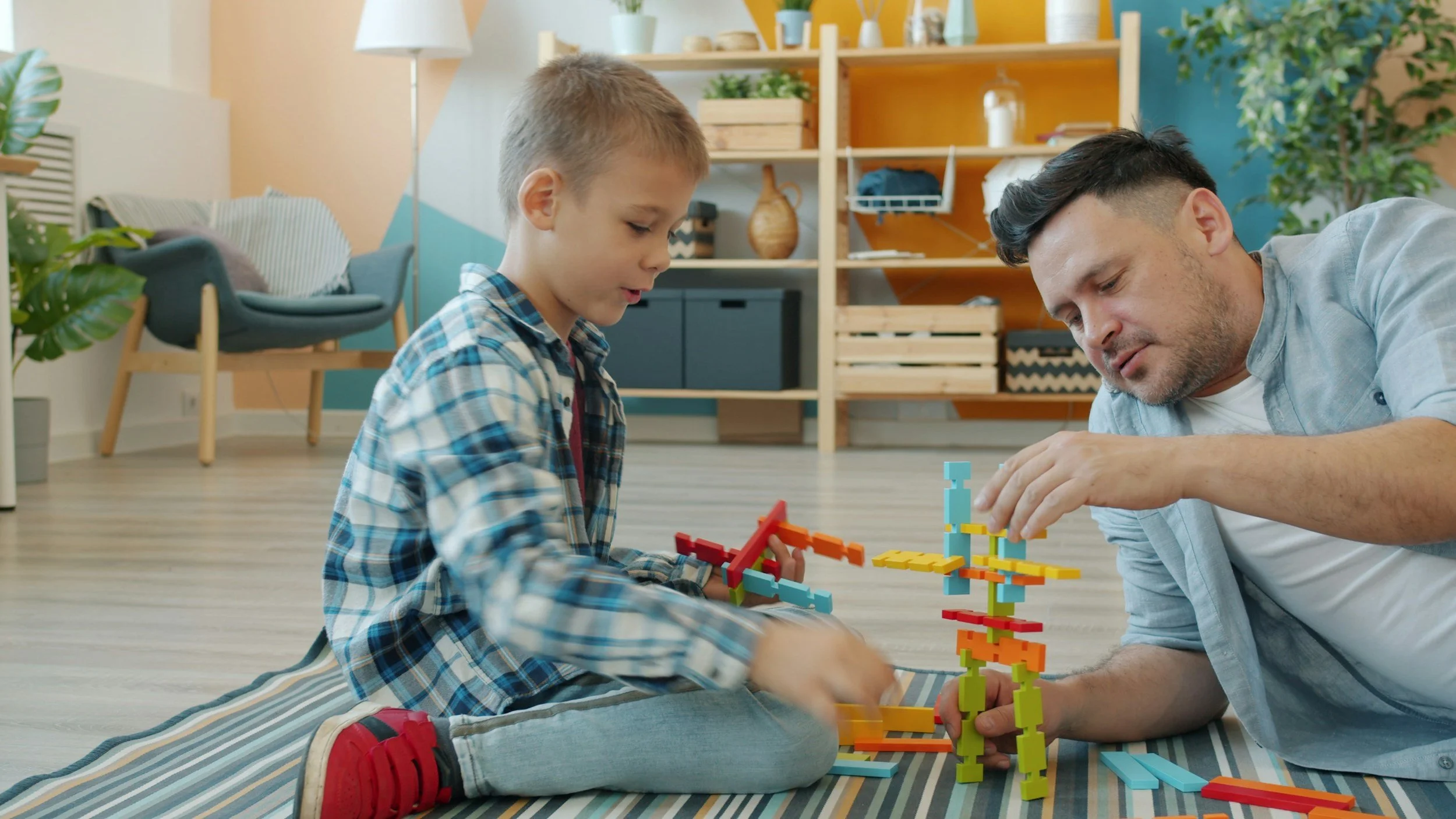 A man and young boy playing with colorful building blocks on a striped rug in a bright living room.