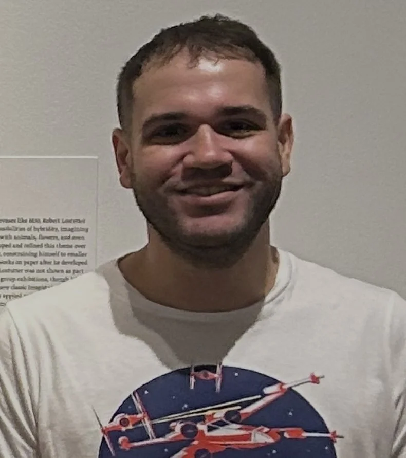 man with short brown hair standing in front of a museum plague and smiling
