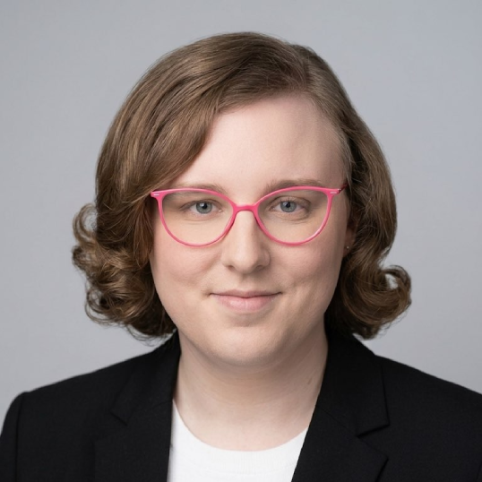 Woman with shoulder length hair and pink glasses, wearing a white shirt and black blazer