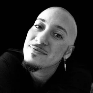 man with goatee, shaved head, and earring, head titled with a closed smile