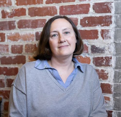 Woman with brown hair just above the chin wearing a blue shirt beneath a v-neck sweater, in front of a brick wall 