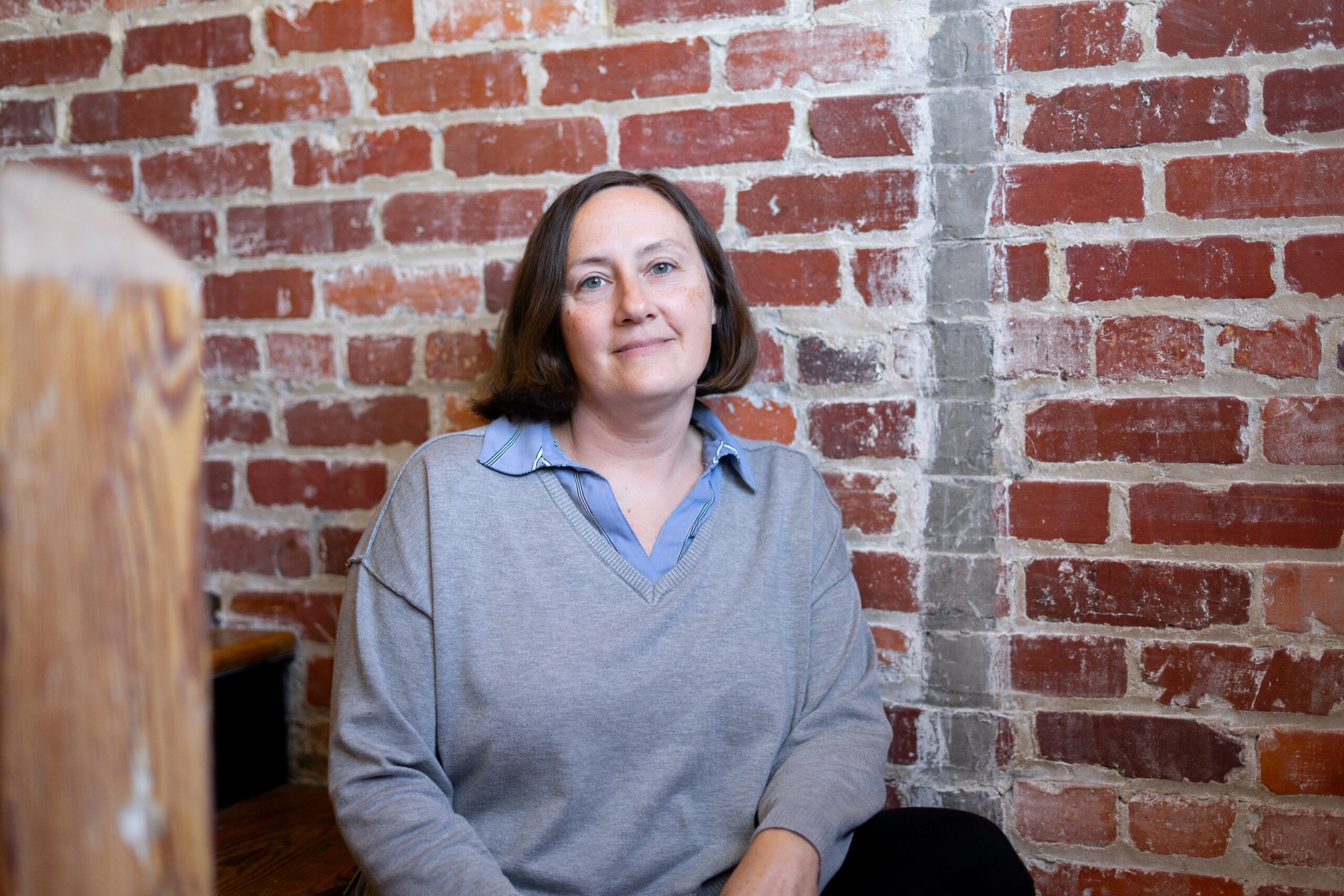 Woman with brown hair just above the chin wearing a blue shirt beneath a v-neck sweater, in front of a brick wall