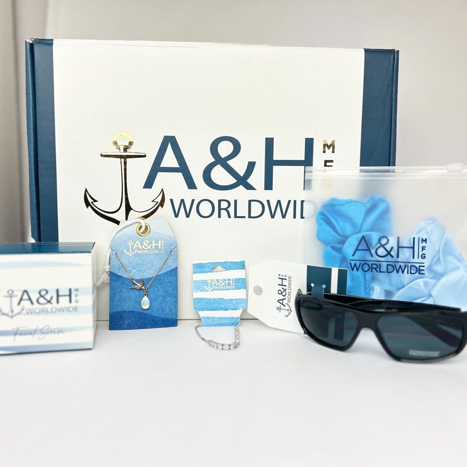 A collection of A&H Worldwide branded items including a box, a necklace, a bracelet, a ring, a pair of sunglasses, a blue scrunchie, and a small box, all arranged on a white surface with a large A&H Worldwide box in the background.