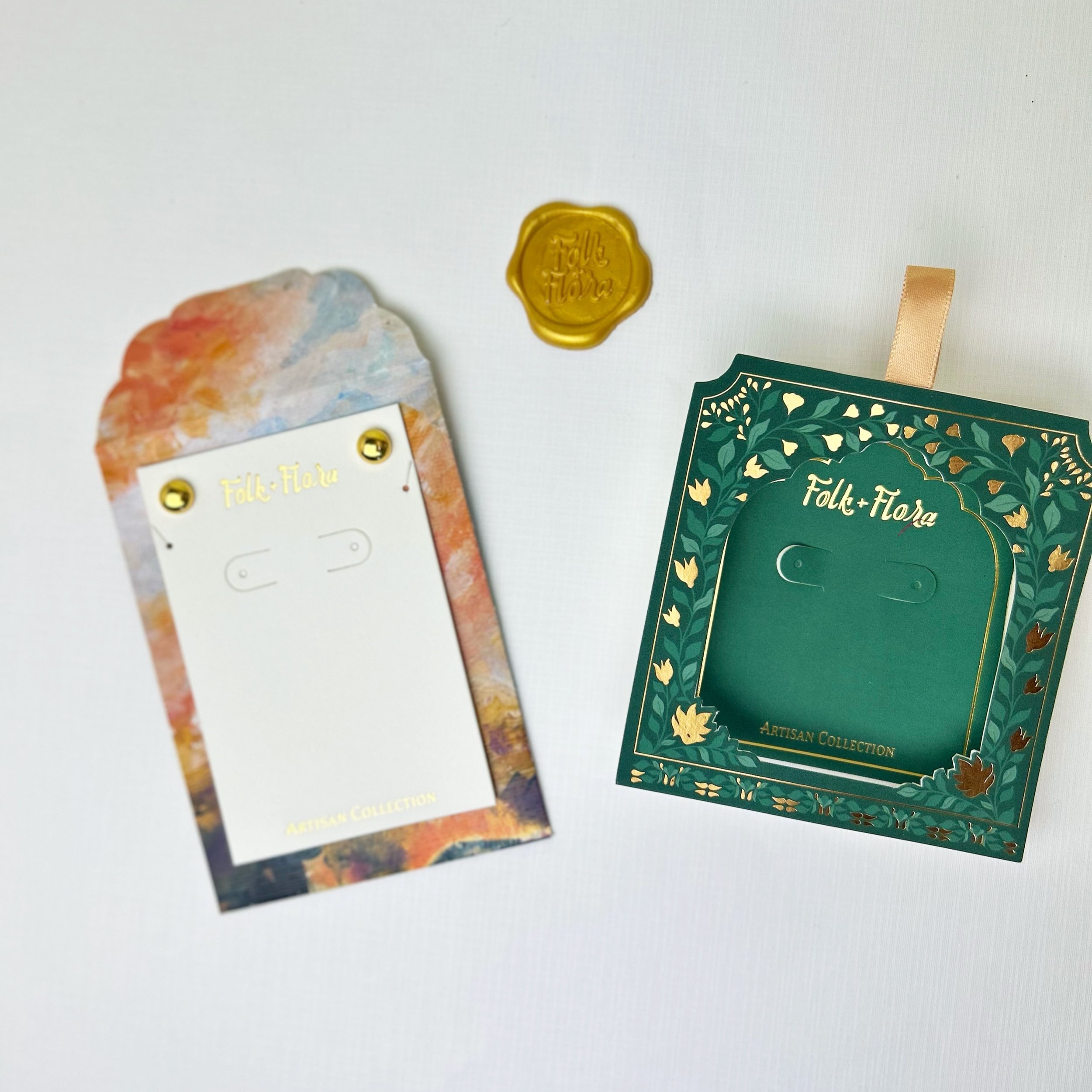 Two decorative cards from the "Folk + Flora" Artisan Collection, one in a colorful abstract design and the other green with gold floral embellishments, along with a gold wax seal.