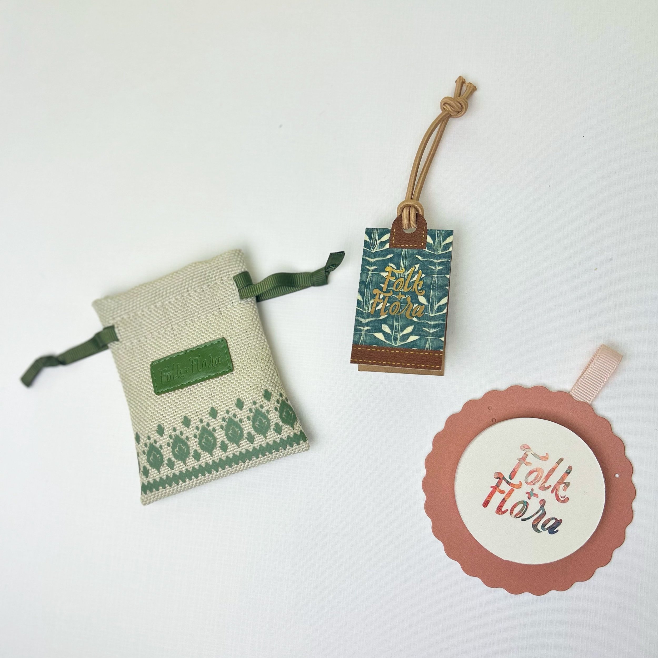Handmade fabric pouch with green leaf design and green drawstring, alongside two decorative tags with "Folk + flora" branding, on a white background.