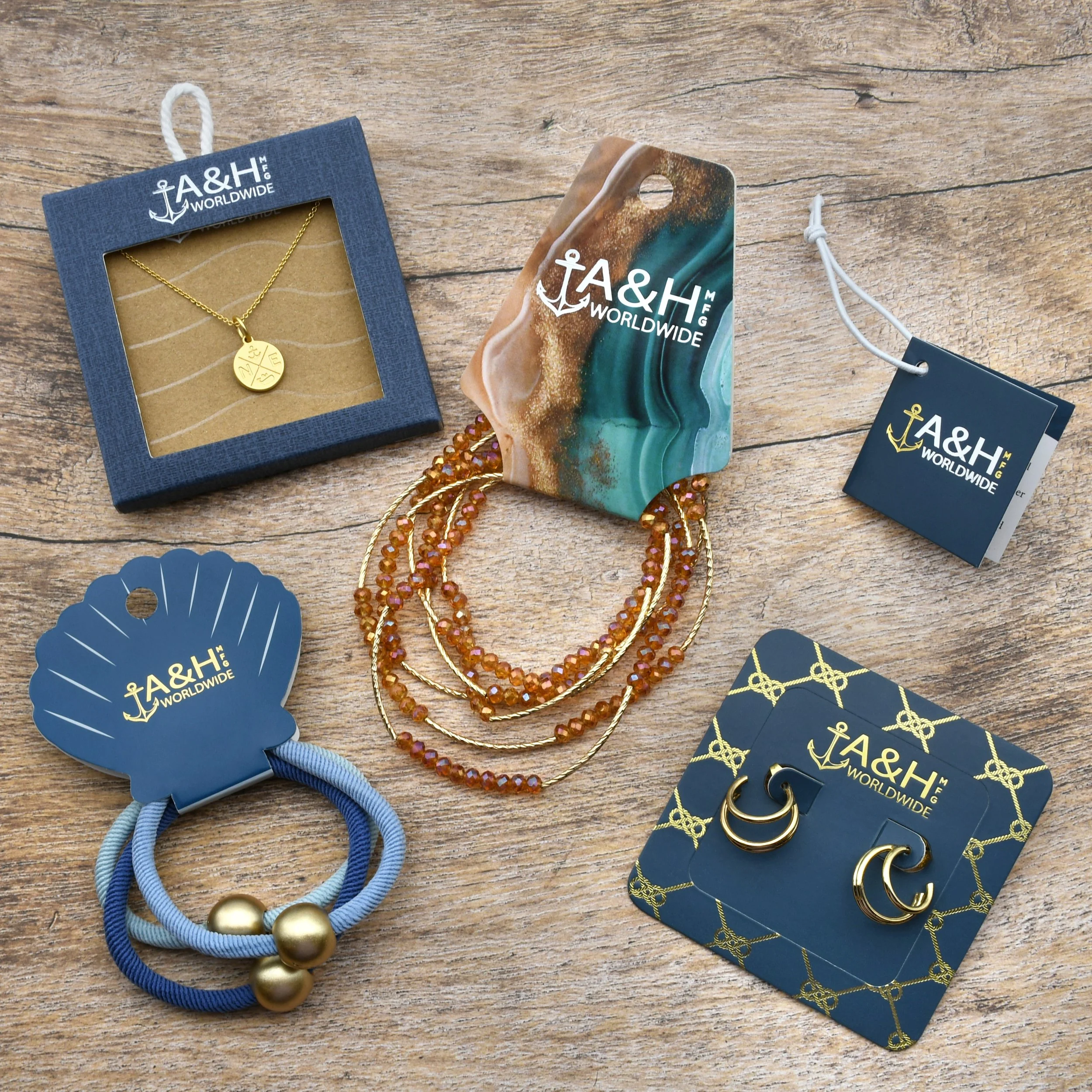 Various jewelry items including a gold necklace with a round pendant, beaded necklaces, and gold hoop earrings, displayed on a wooden surface with branded tags and packaging from A&H Worldwide.