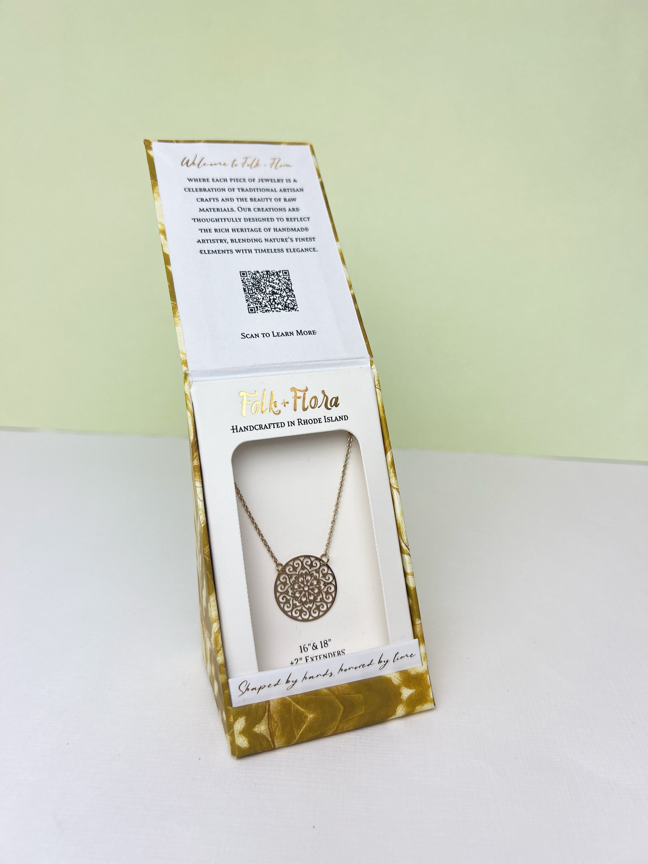 Decorative necklace in box with intricate circular pendant and gold chain, featuring Folk + Flora branding and packaging details.