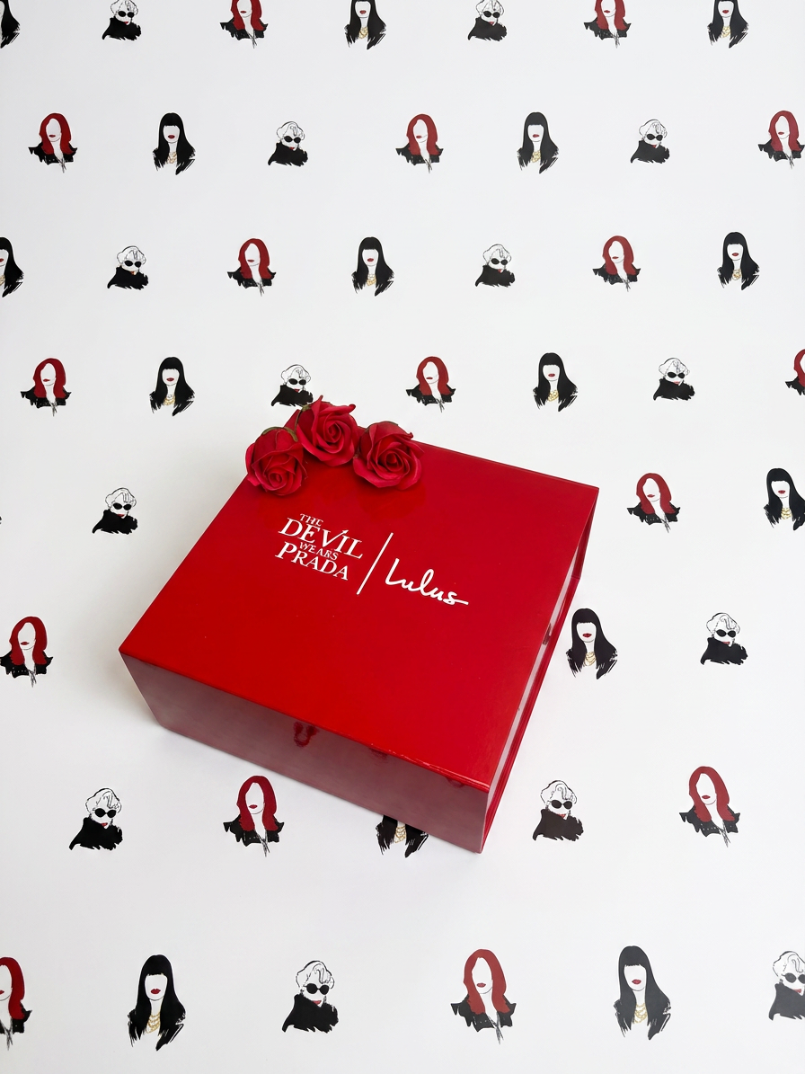 A red box titled 'The Devil Wears Prada | Lula's' with three red roses on top, set against a white background featuring a repeating pattern of illustrated women with black hair, red hair, and sunglasses. influencer bespoke boxes packaging