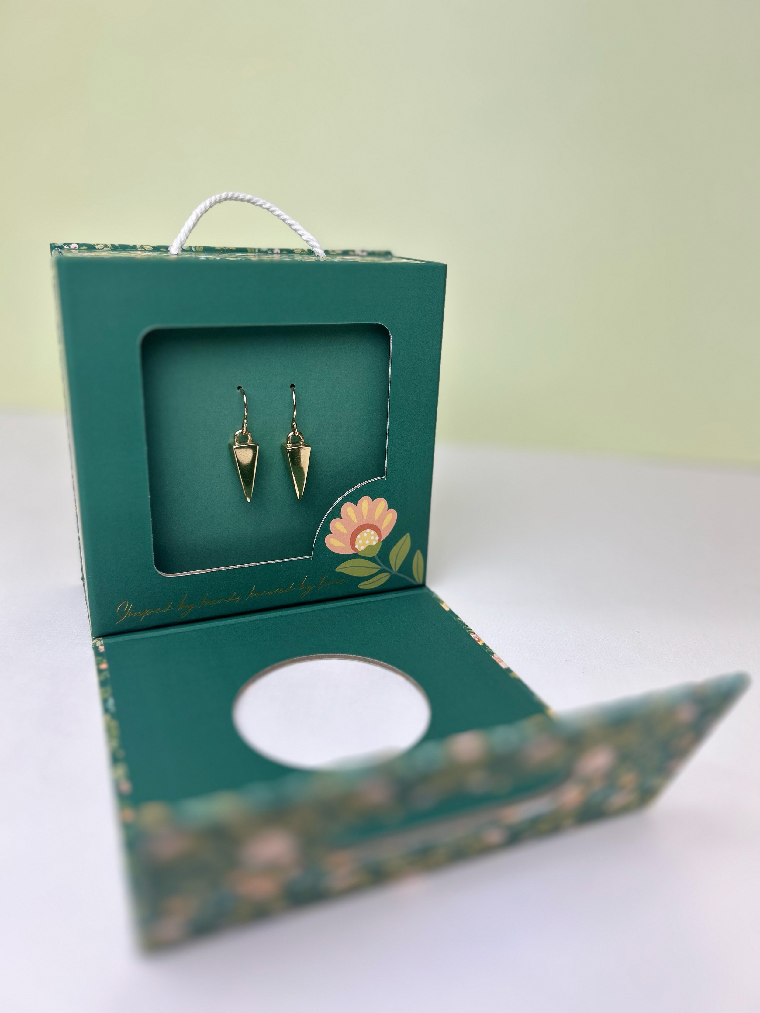 A pair of gold earrings displayed inside an open green box with floral design and a handle.