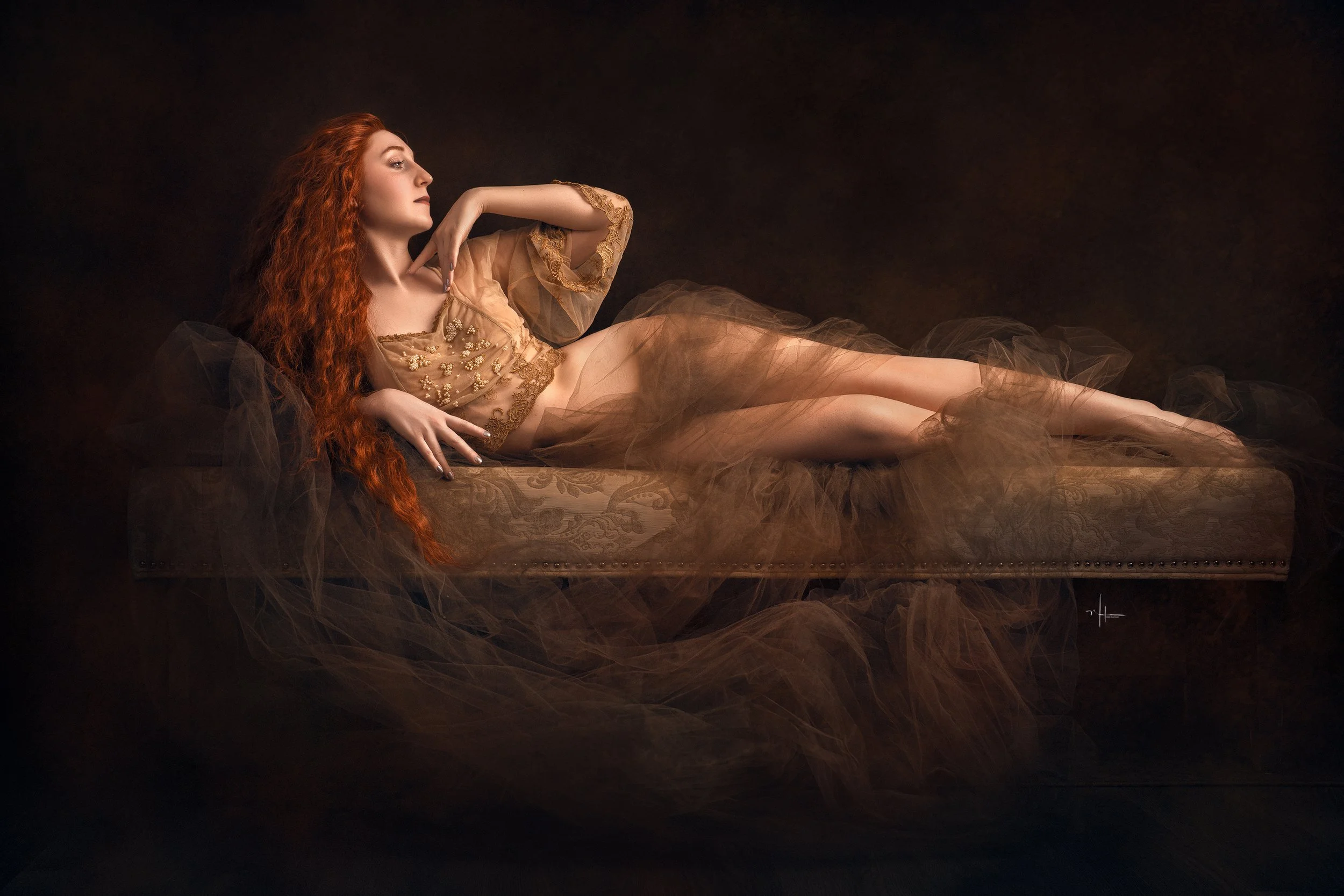 A woman with long red hair lying on a decorated sofa, dressed in a sheer beige gown with embroidery, against a dark background.