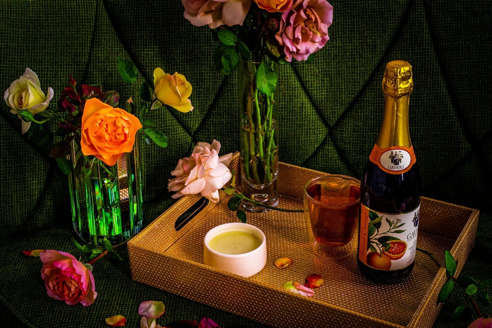 A decorative scene with pink, orange, yellow, and white roses in vases, a bottle of Gavioli sparkling wine, a glass of rosé wine, a small bowl of green dip, and scattered rose petals on a golden tray against a green textured fabric background.