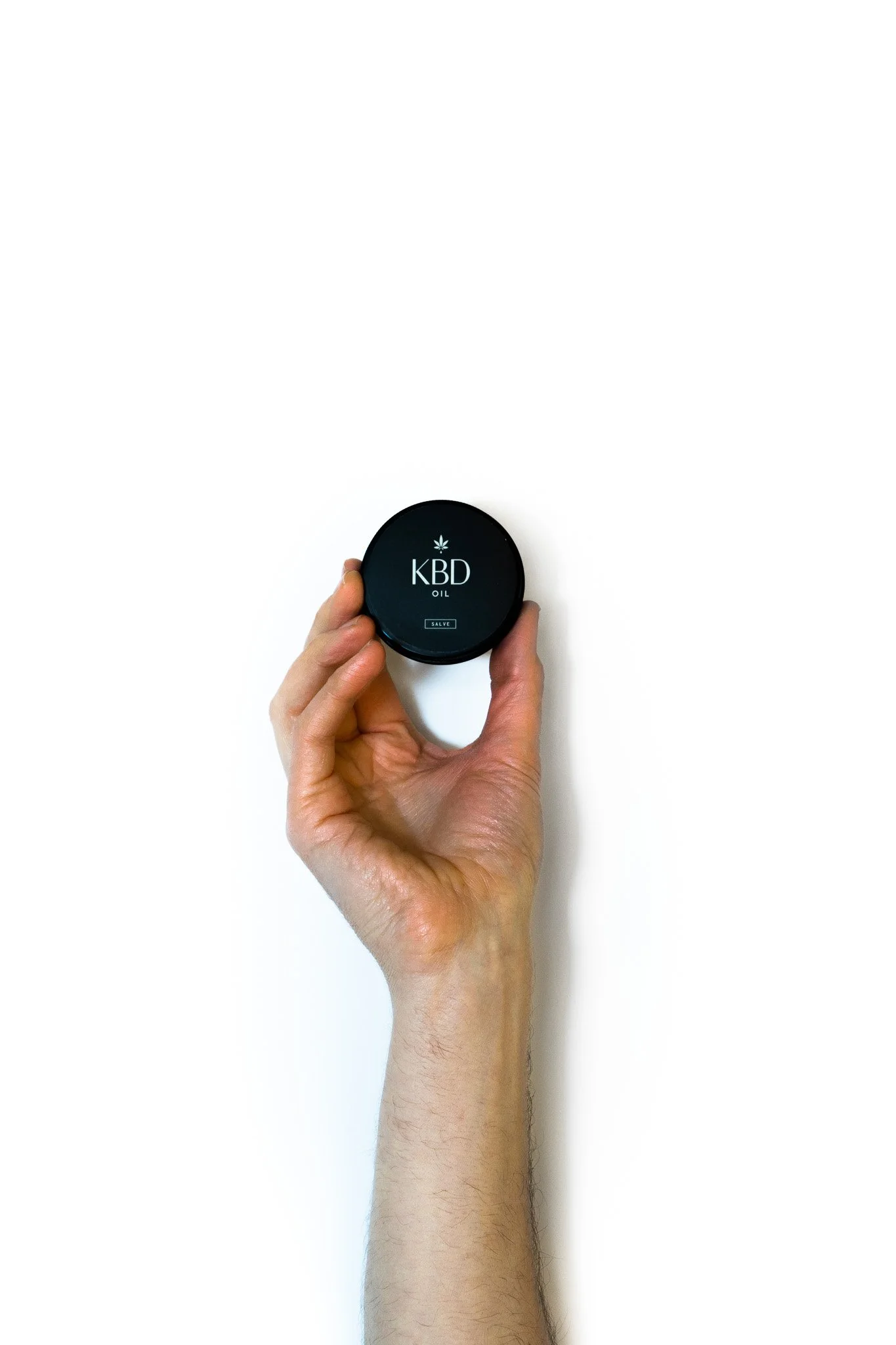 Hand holding a small black container labeled 'KBD Oil' with a cannabis leaf symbol on a plain white background.