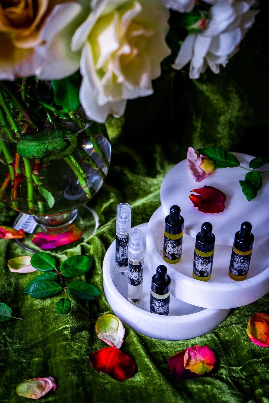 A display of skincare serums in small bottles surrounded by green leaves, rose petals, and a white orchid, arranged on a green velvet fabric with a white marble tray.