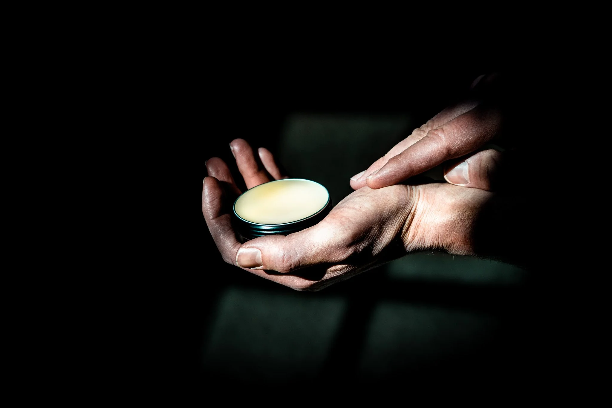 A person holding an open container of white cream or lotion in one hand, while the other hand is applying the cream to their skin, with a dark background.