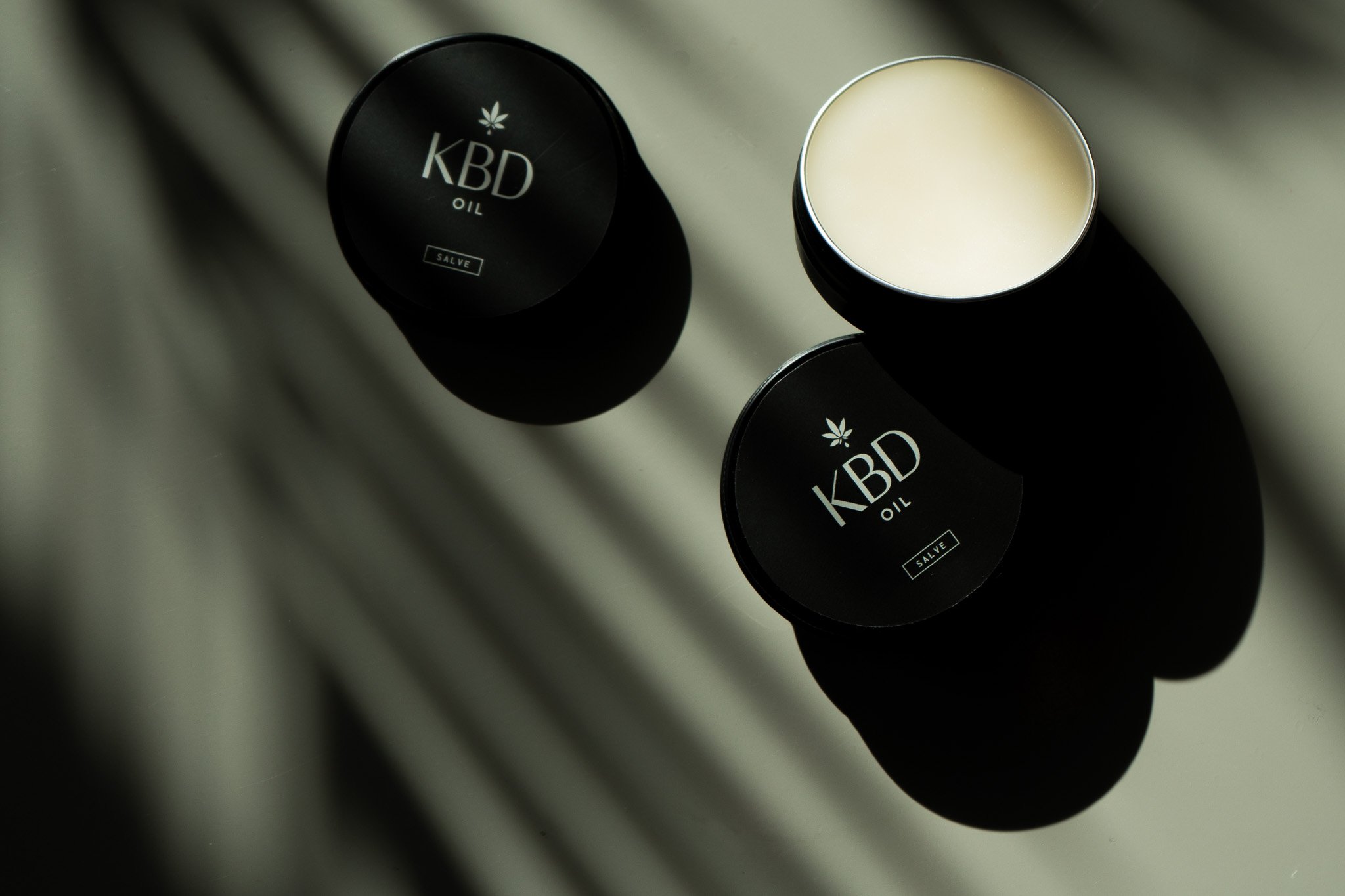 Two black containers labeled 'KBD Oil' with one open, revealing a cream-colored substance inside, placed on a reflective surface.