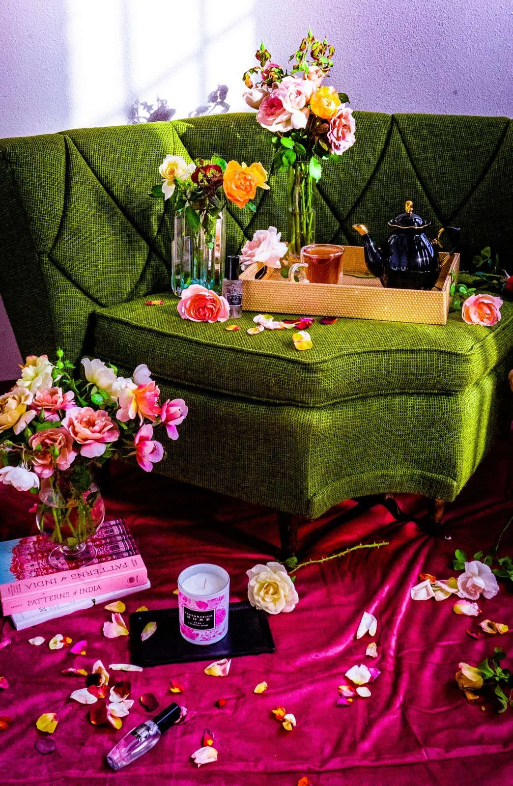 A green vintage sofa with floral arrangements on top, a black teapot, a mug, and a tray. The sofa is on a pink cloth decorated with scattered flower petals, candles, books, and small items.