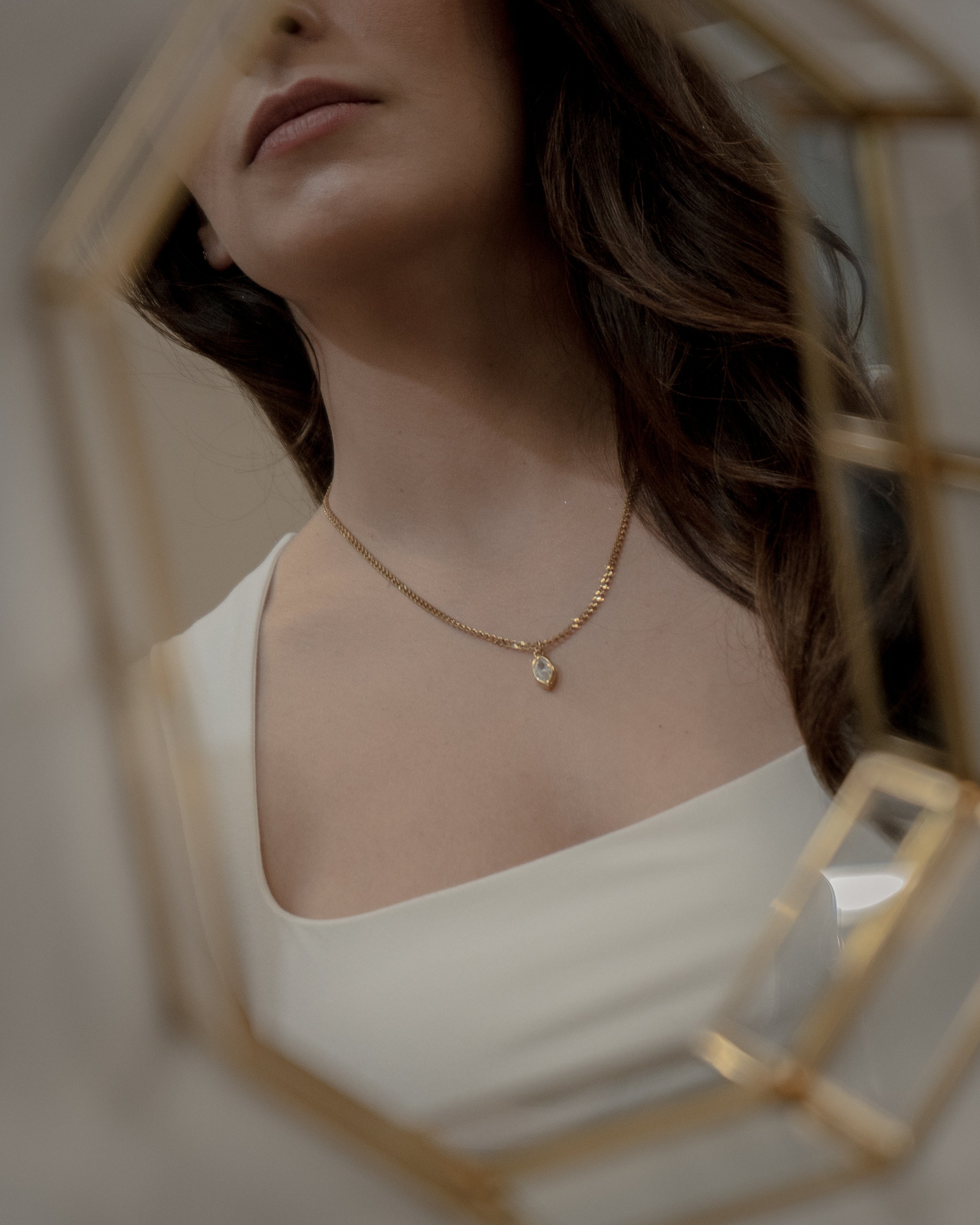 Close-up of a woman’s neck and chin, wearing a gold necklace with a small pendant, reflected in a mirror with a gold frame.