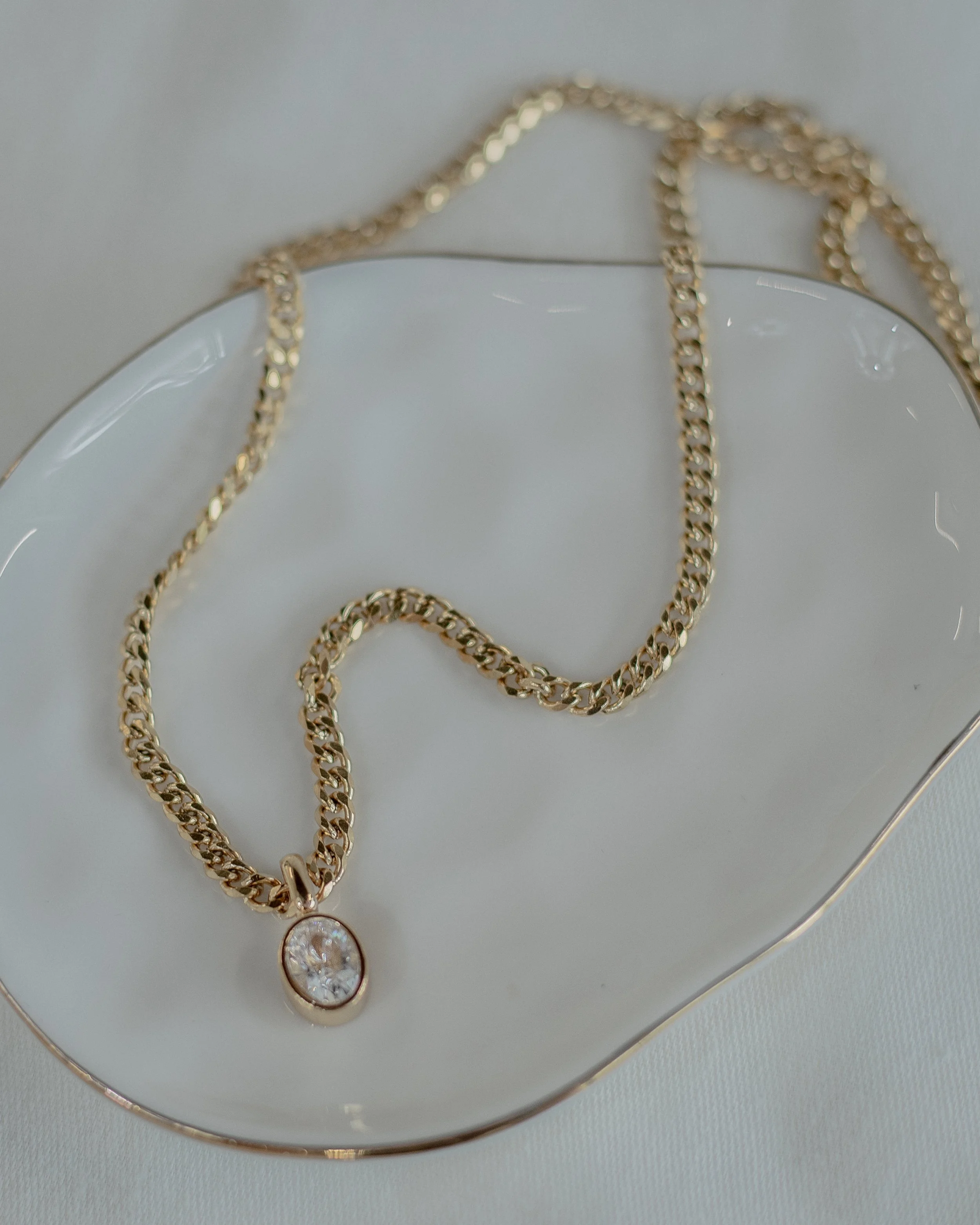 Gold necklace with a diamond pendant in a gold bezel, resting on a white, wavy-edged plate.