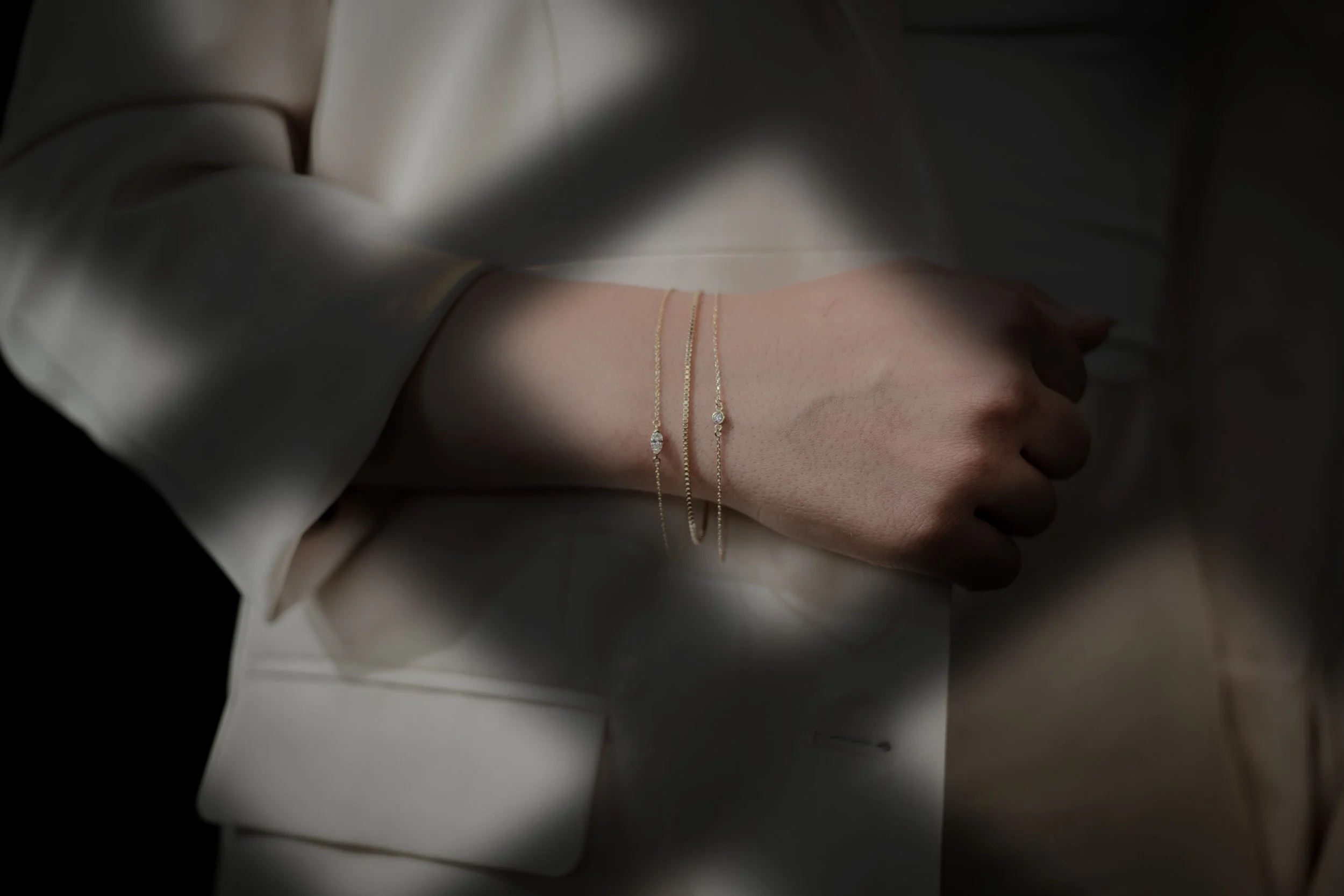 Close-up of a woman's hand wearing two delicate gold bracelets with small diamonds, partially covered by the sleeve of a cream-colored satin blazer.