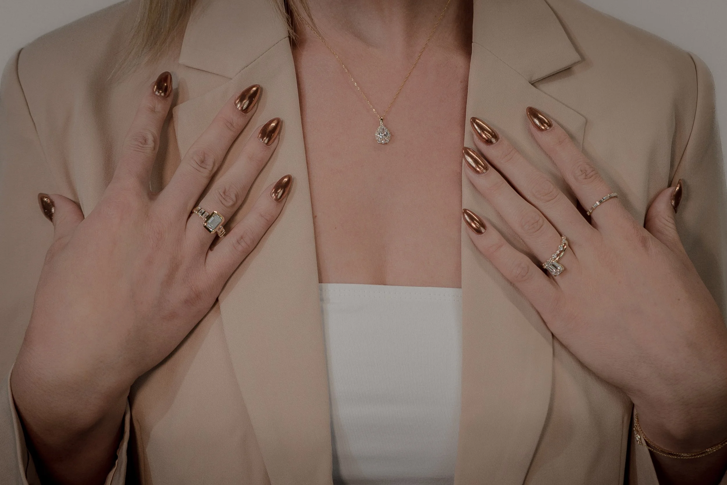 Close-up of a woman wearing jewelry, with fingers on her chest, dressed in a beige blazer and a white top.