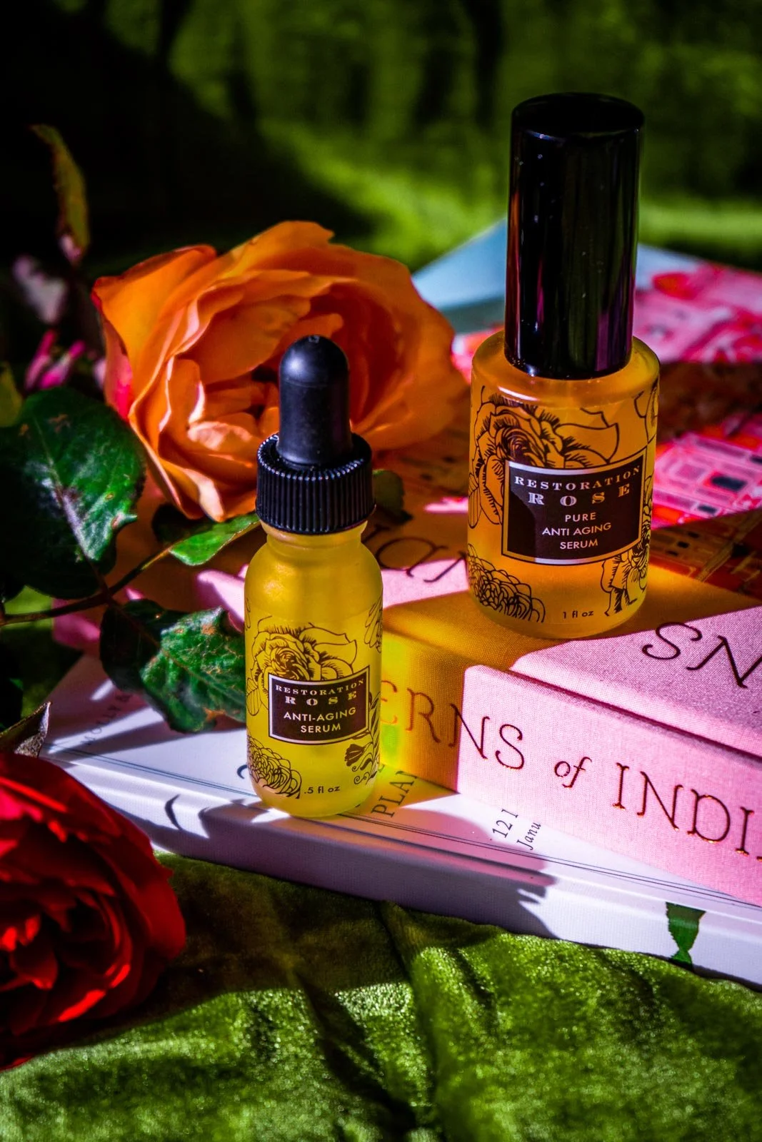 Two bottles of anti-aging serum labeled Restoration Rose placed on top of books, surrounded by roses and greenery, with a blurred outdoor background.