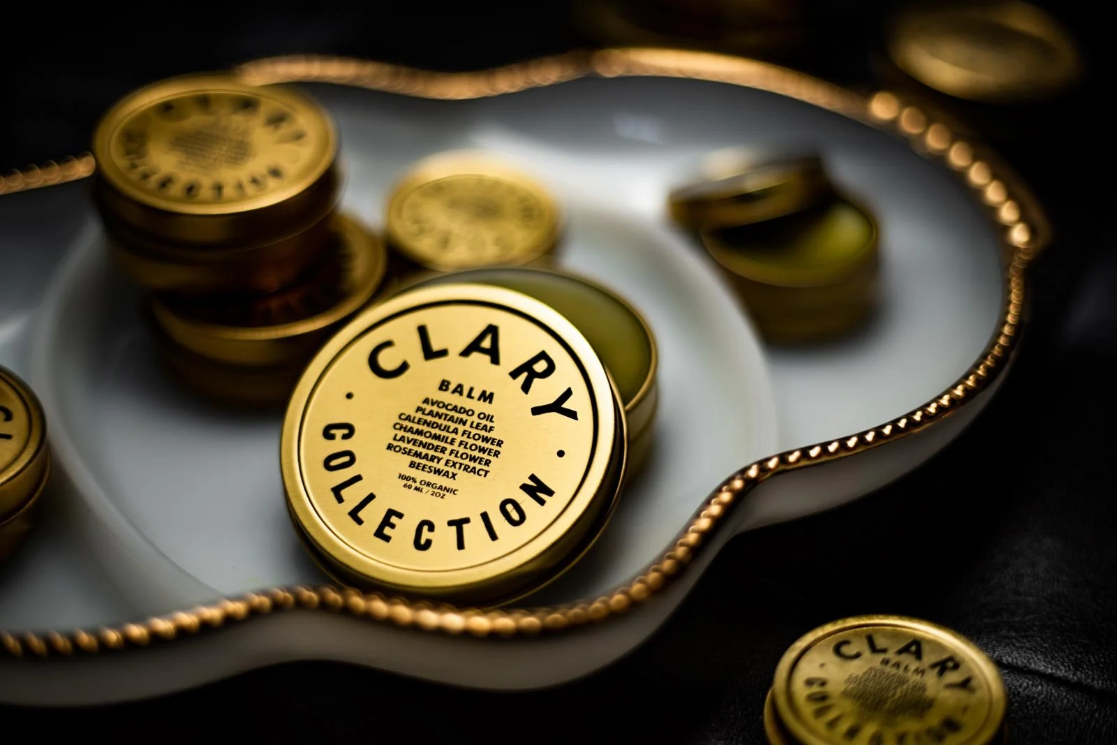 A tin labeled 'Clear Collection' containing a skincare balm with ingredients listed, surrounded by other tins on a reflective surface with a decorative gold-trimmed mirror.
