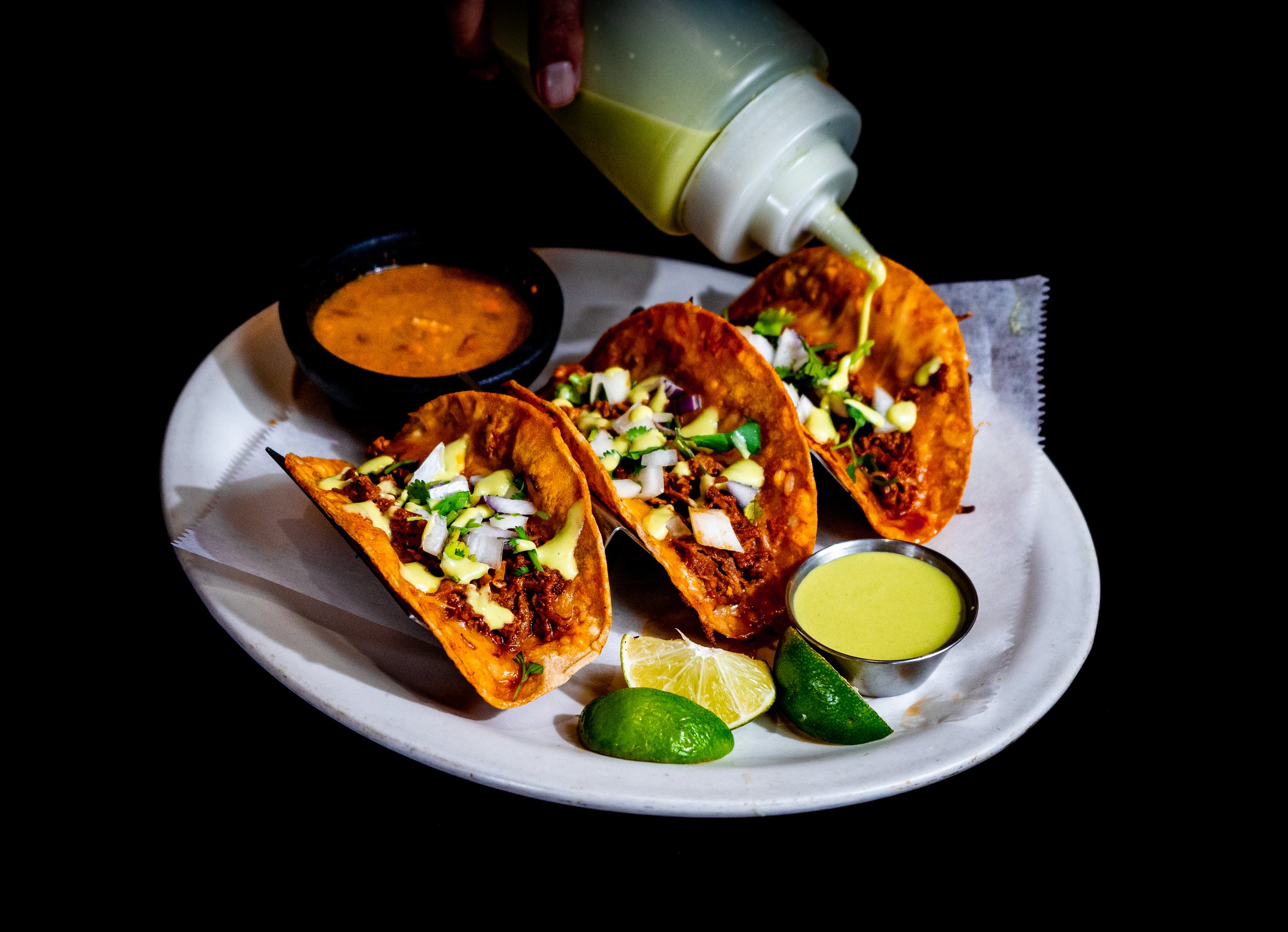 Three tacos with chopped onions and cilantro, two lime wedges, and two small bowls of sauce on a white plate, with a squeeze bottle of green sauce pouring onto the tacos.