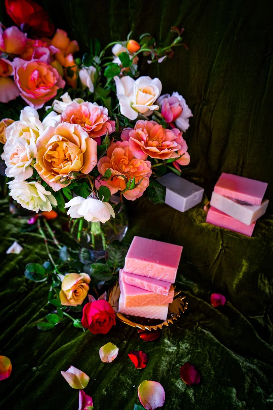 A bouquet of pink, white, and peach roses with soap bars on a dark green cloth and a wooden surface.