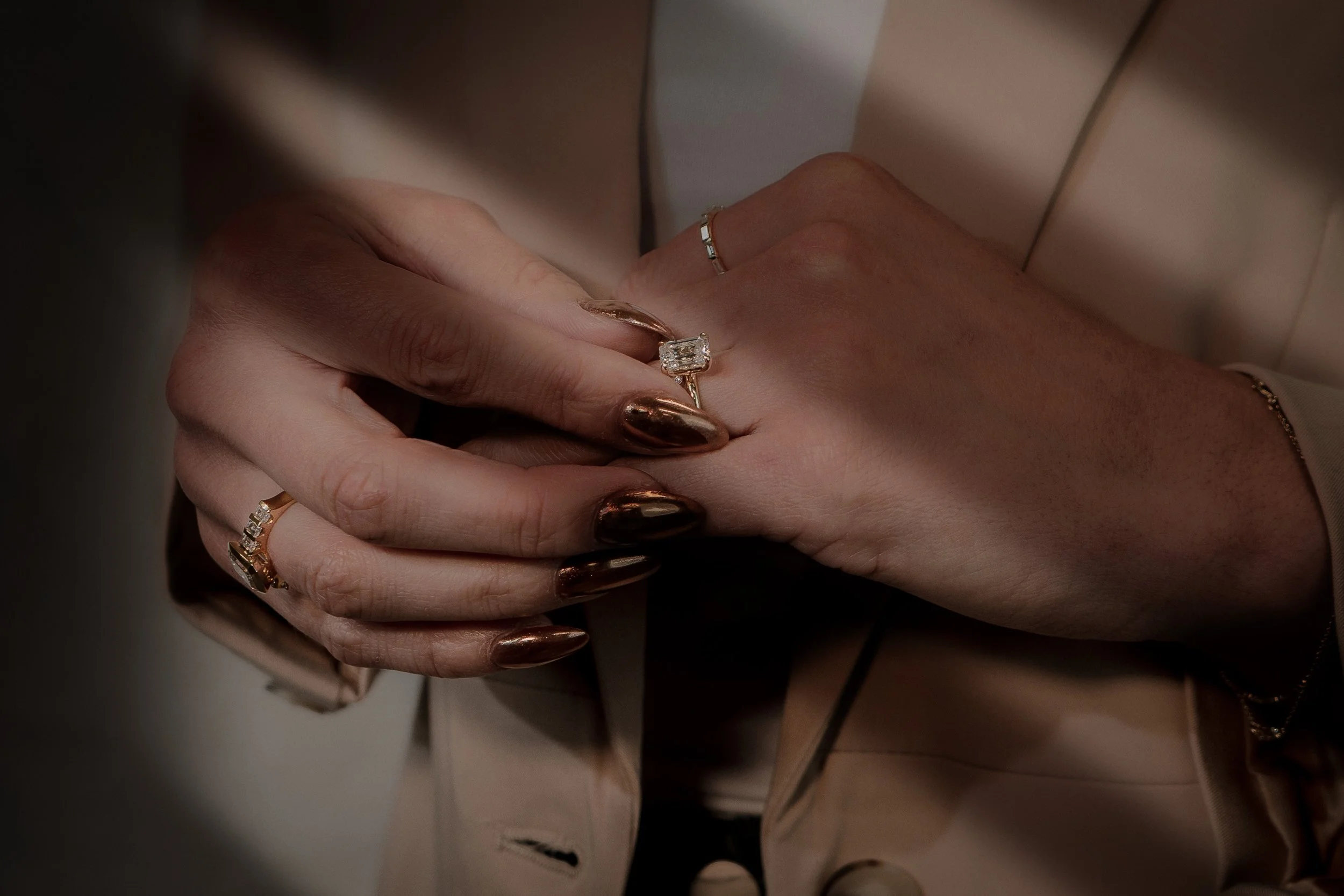 Close-up of a woman’s hands adorned with rings, including a large diamond ring, and metallic nail polish. The woman is wearing a beige blazer.
