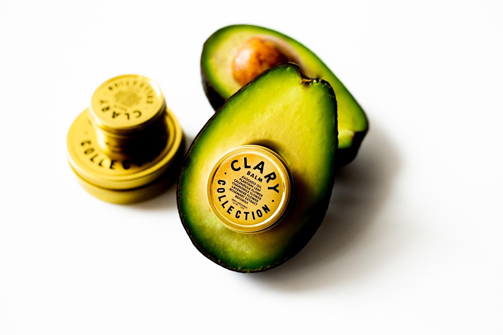 A halved avocado with a Clary Collection balm container placed on top, and a cluster of small balm containers in the background on a white surface.