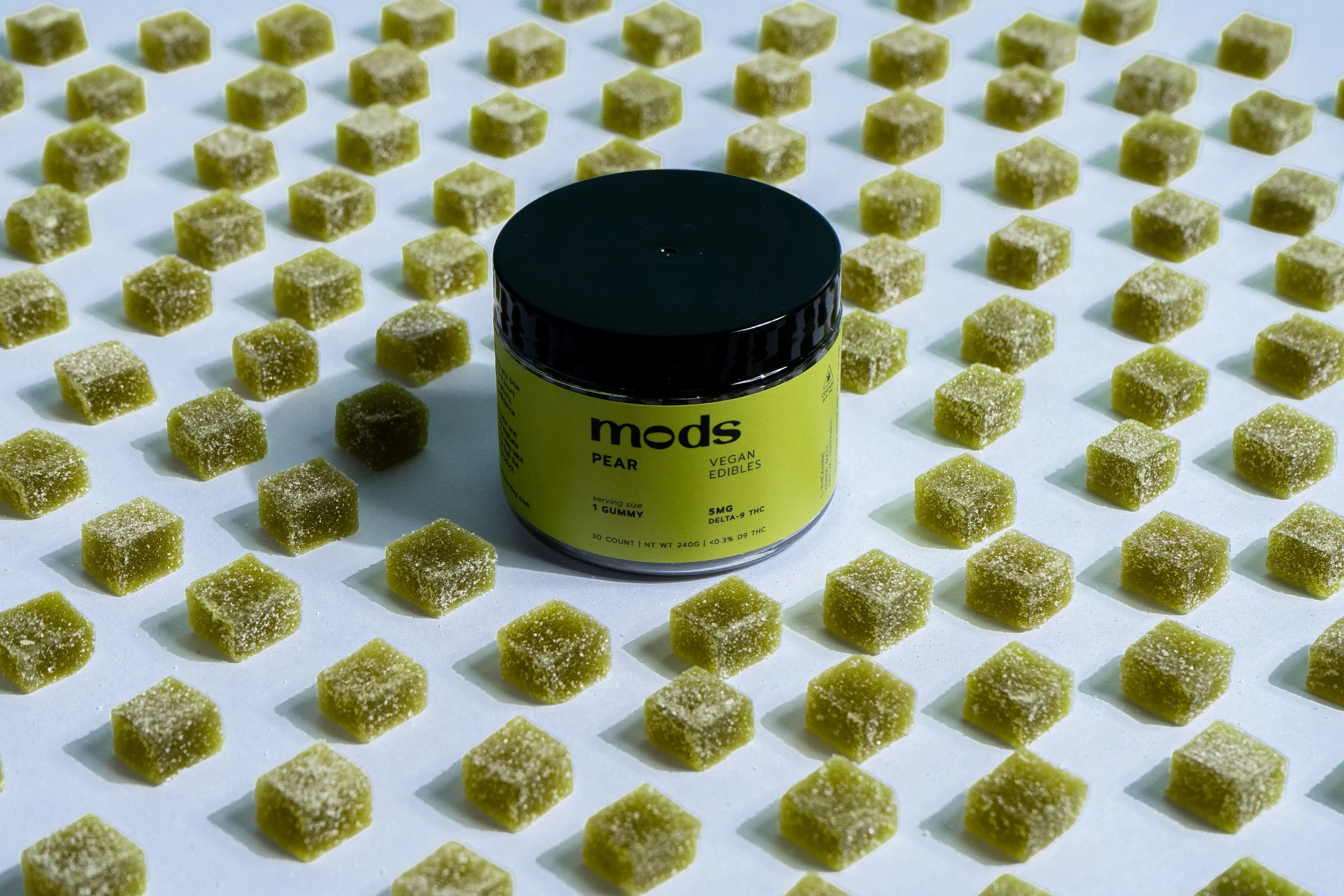 Container of vegan pear edibles surrounded by numerous green square gummies coated in granulated sugar, arranged in rows on a white surface.