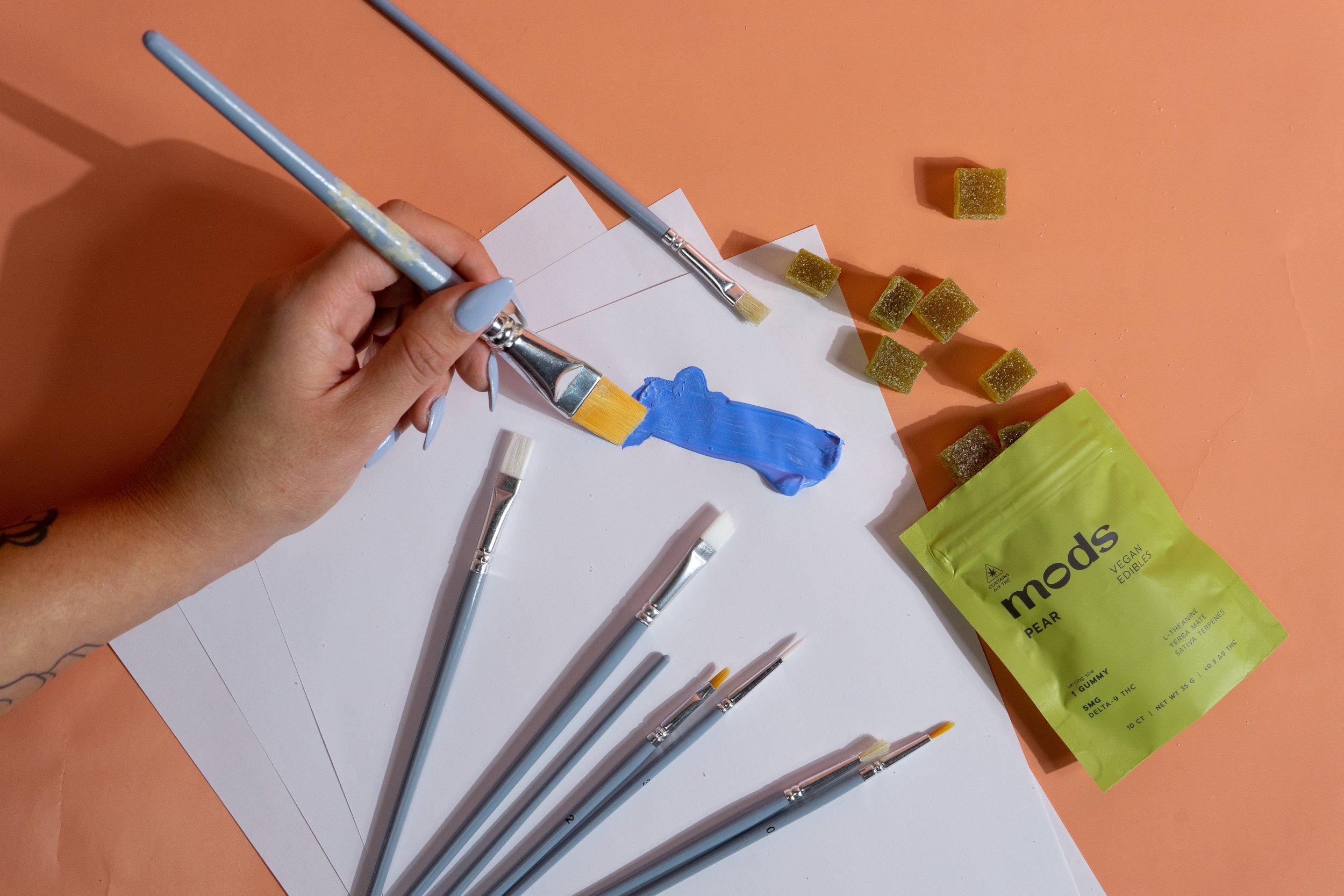 A person painting a strip of blue paint on white paper with a small paintbrush, with several other paintbrushes and gummy candies on an orange surface.