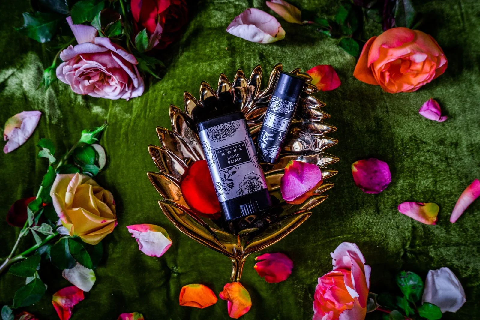 Two rose-scented products on a decorative dish, surrounded by various scattered rose petals and flowers on a green fabric background.