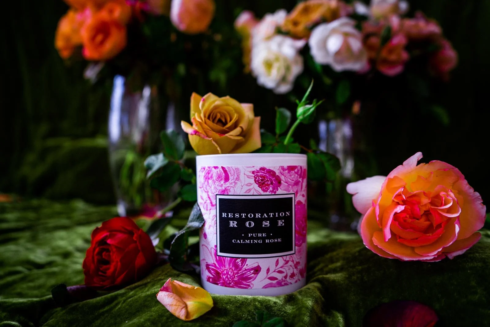 A pink and white floral labeled candle on a green velvet surface surrounded by colorful roses and flower vases.