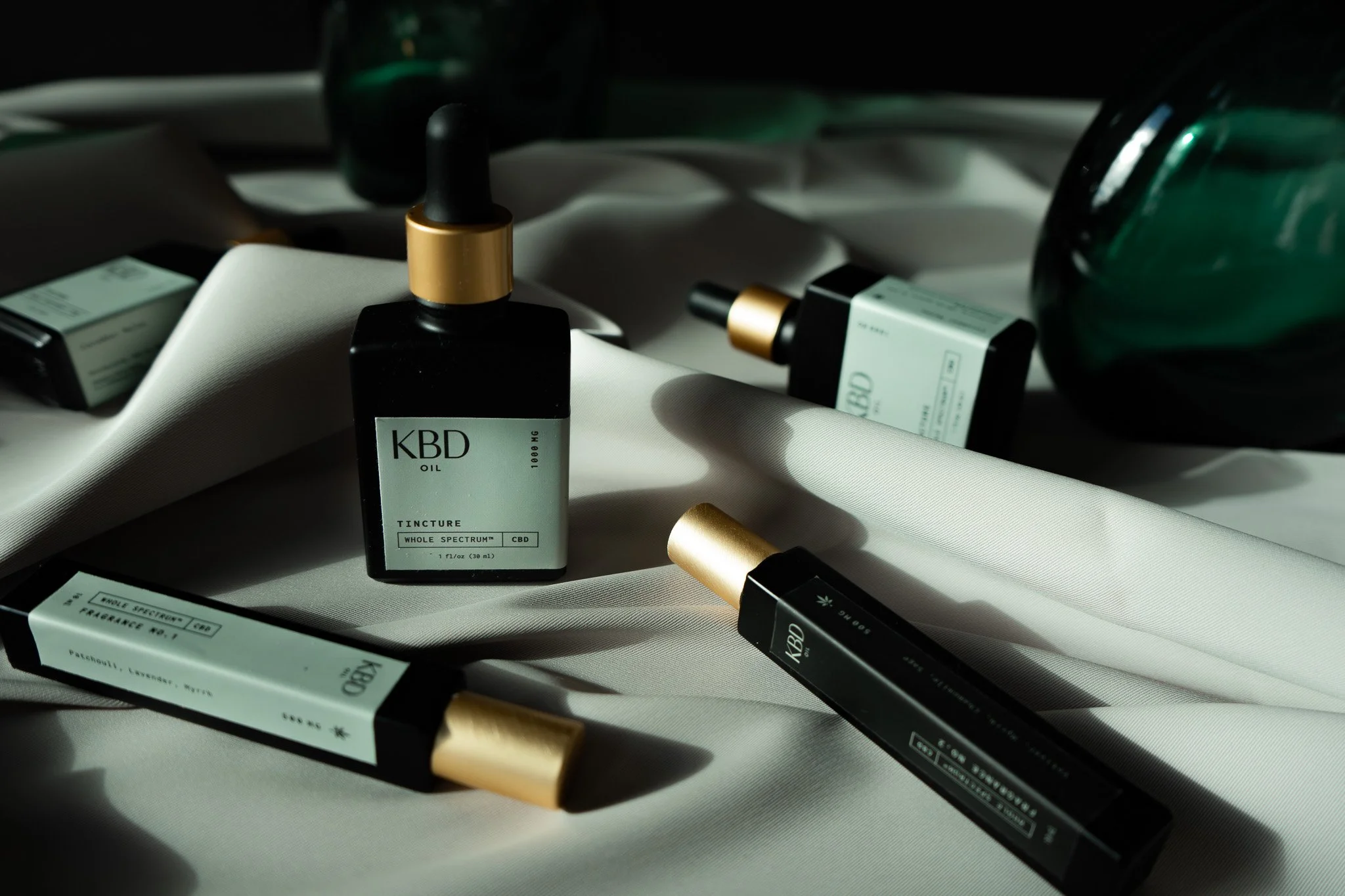 Several KBD CBD oil bottles and vape cartridges scattered on a white fabric surface with shadows and reflections.