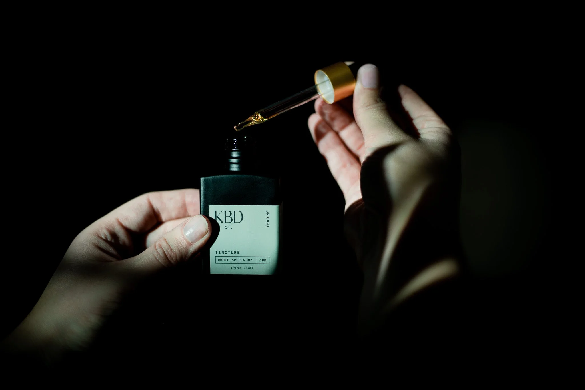 Hands holding a small dropper bottle of CBD oil with a dropper above it, against a black background.
