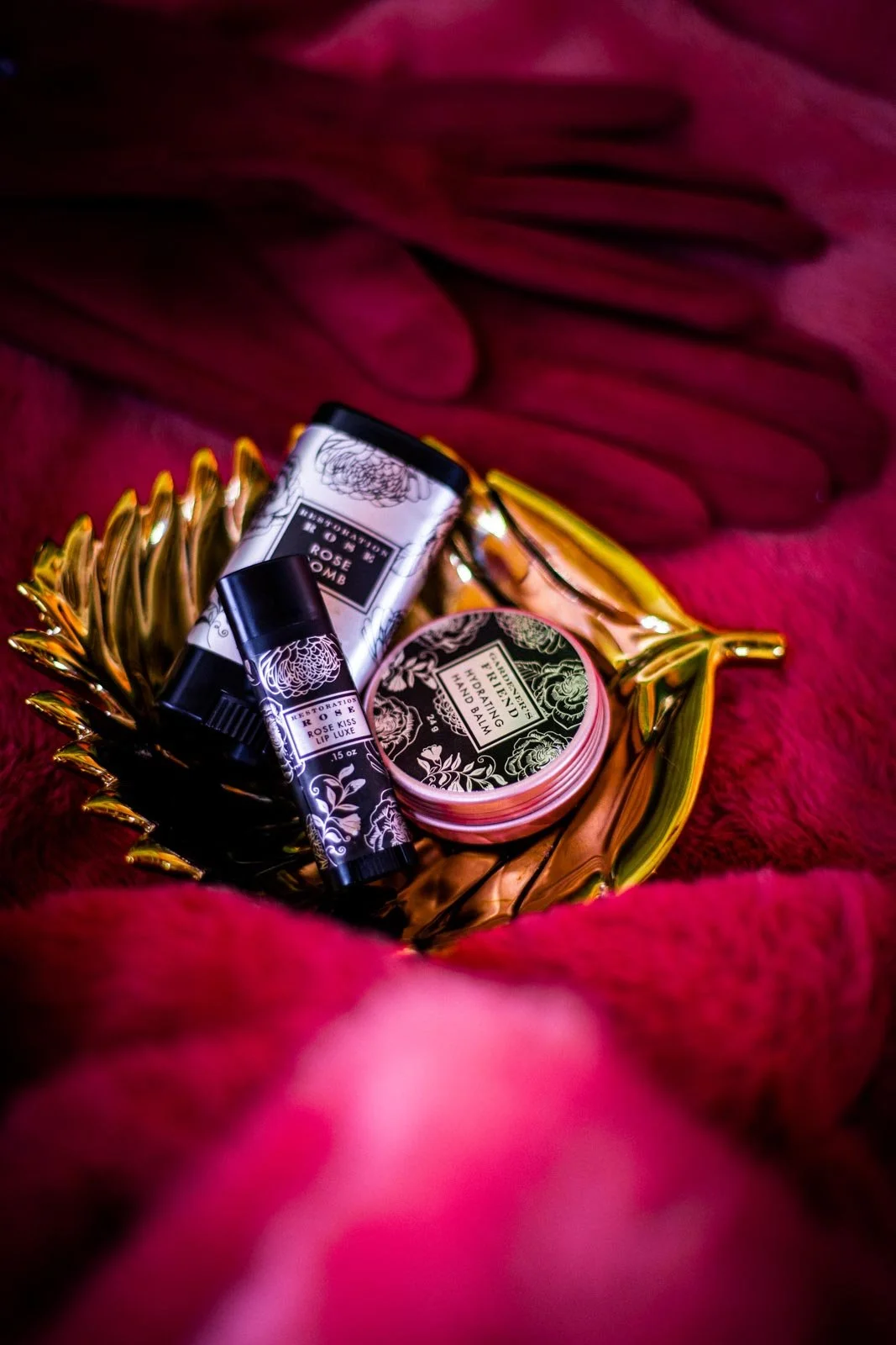 A gold decorative bowl filled with small skincare products including a rose-scented lipstick, a tube of lip balm, and a jar of hydrating hand balm, placed on a plush red fabric background.