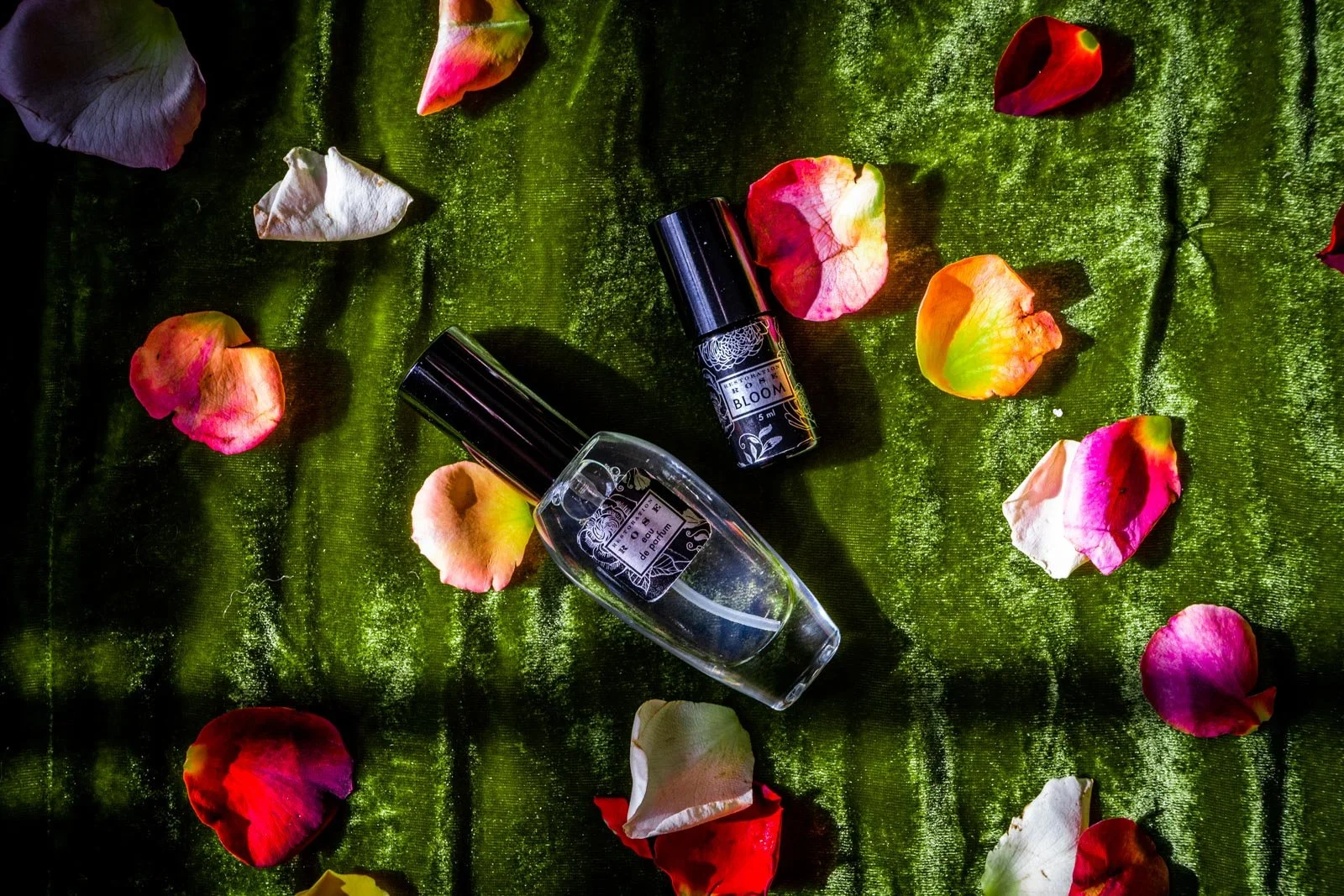A perfume bottle and a small spray bottle are placed on a green velvet cloth, surrounded by scattered rose petals in shades of pink, white, yellow, and red.
