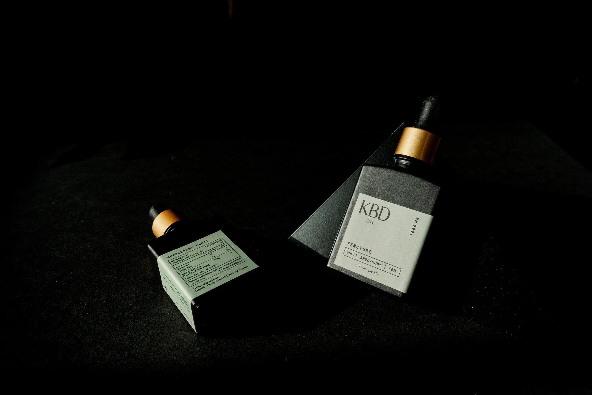Two black bottles of CBD oil, one in a box and one lying next to it, on a dark surface.