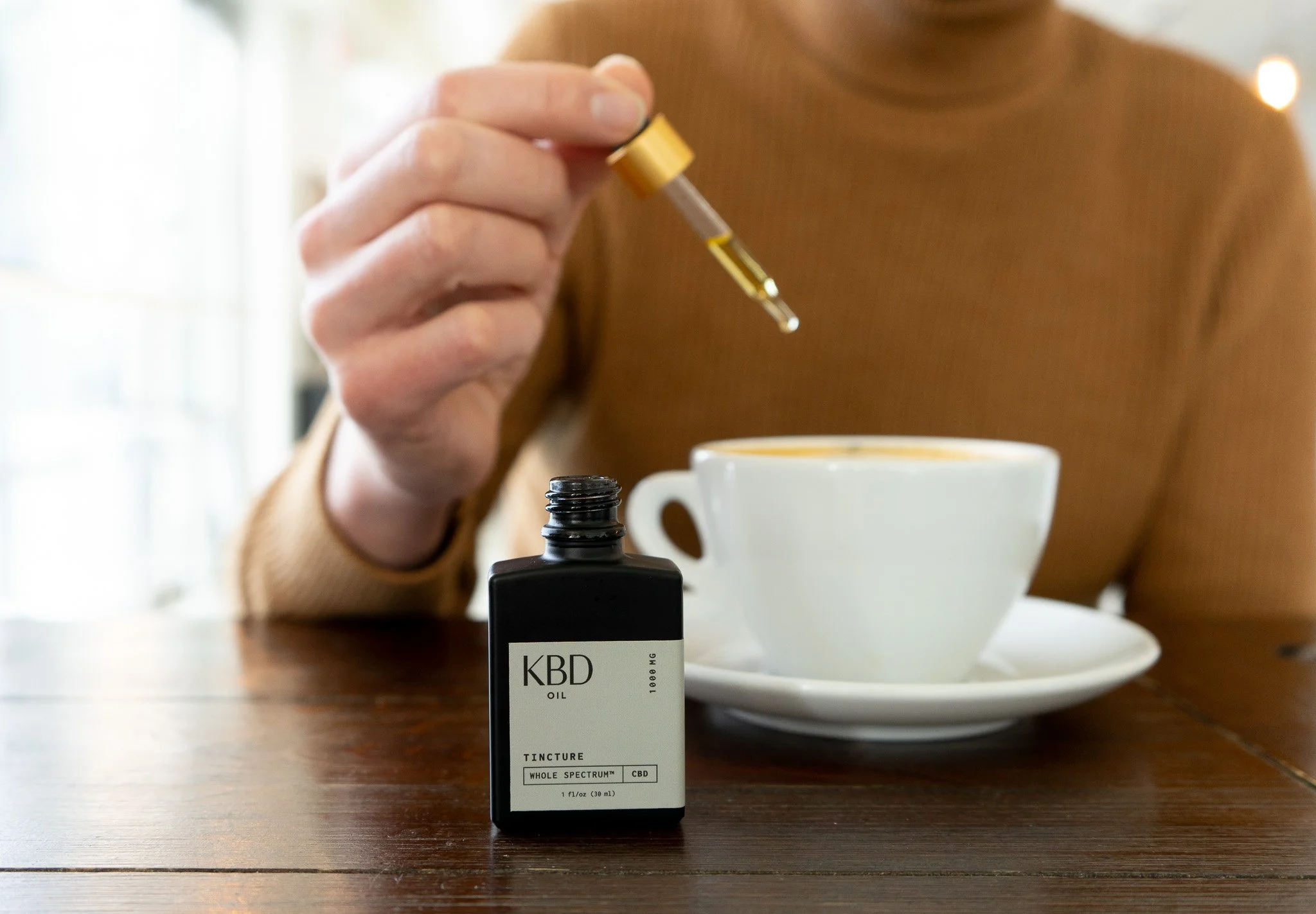 A person holding a dropper with CBD oil over a white coffee cup on a wooden table, with a black bottle of CBD oil labeled 'KBD' in the foreground.