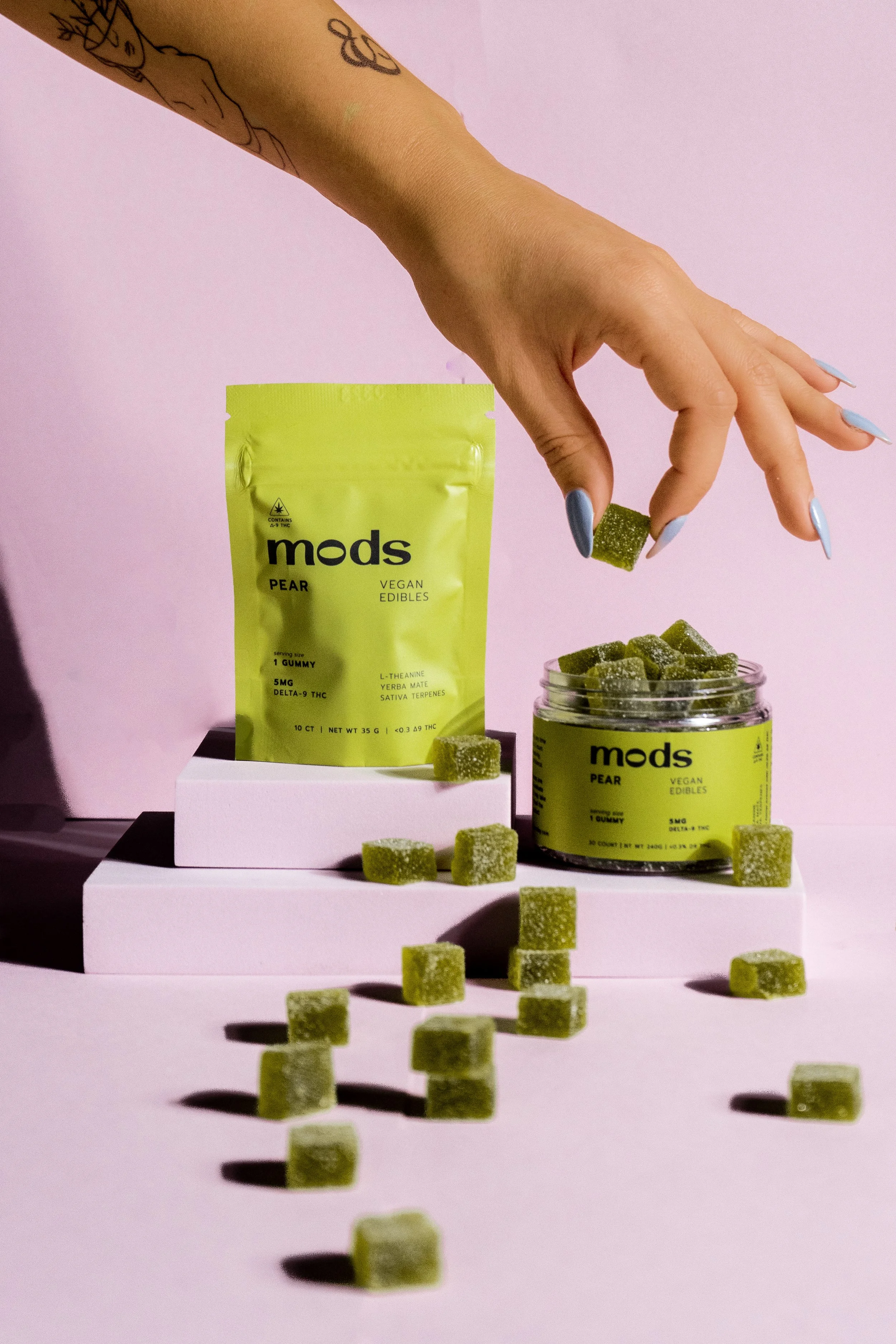 A person with blue painted fingernails holding a green gummy candy over a jar of similar candies, with a pink background and a packaging pouch of 'Mods Pear Vegan Edibles' and additional candies on display.