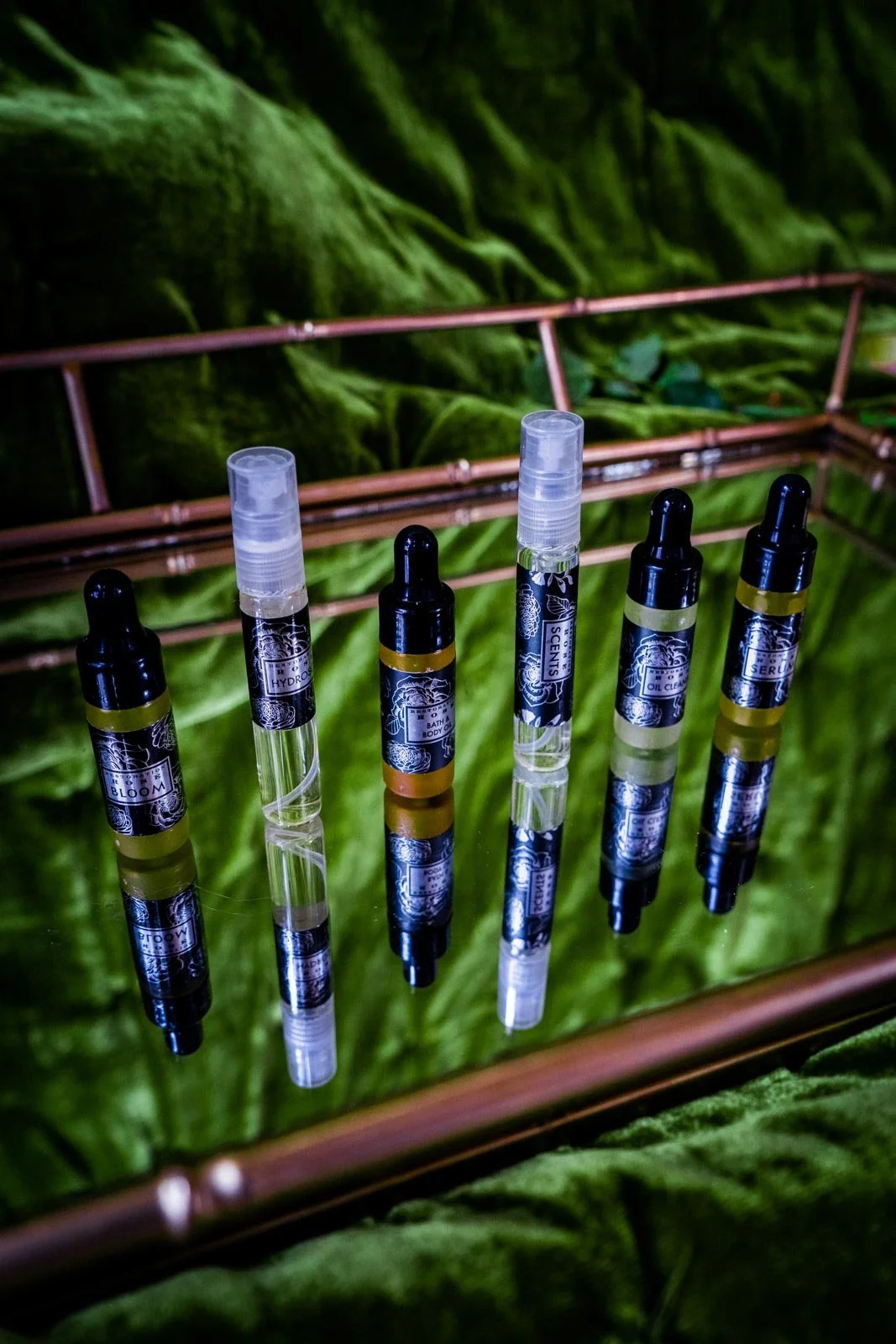 Six small cosmetic spray bottles with black labels on a reflective surface, set against a green fabric backdrop with a copper-colored frame.