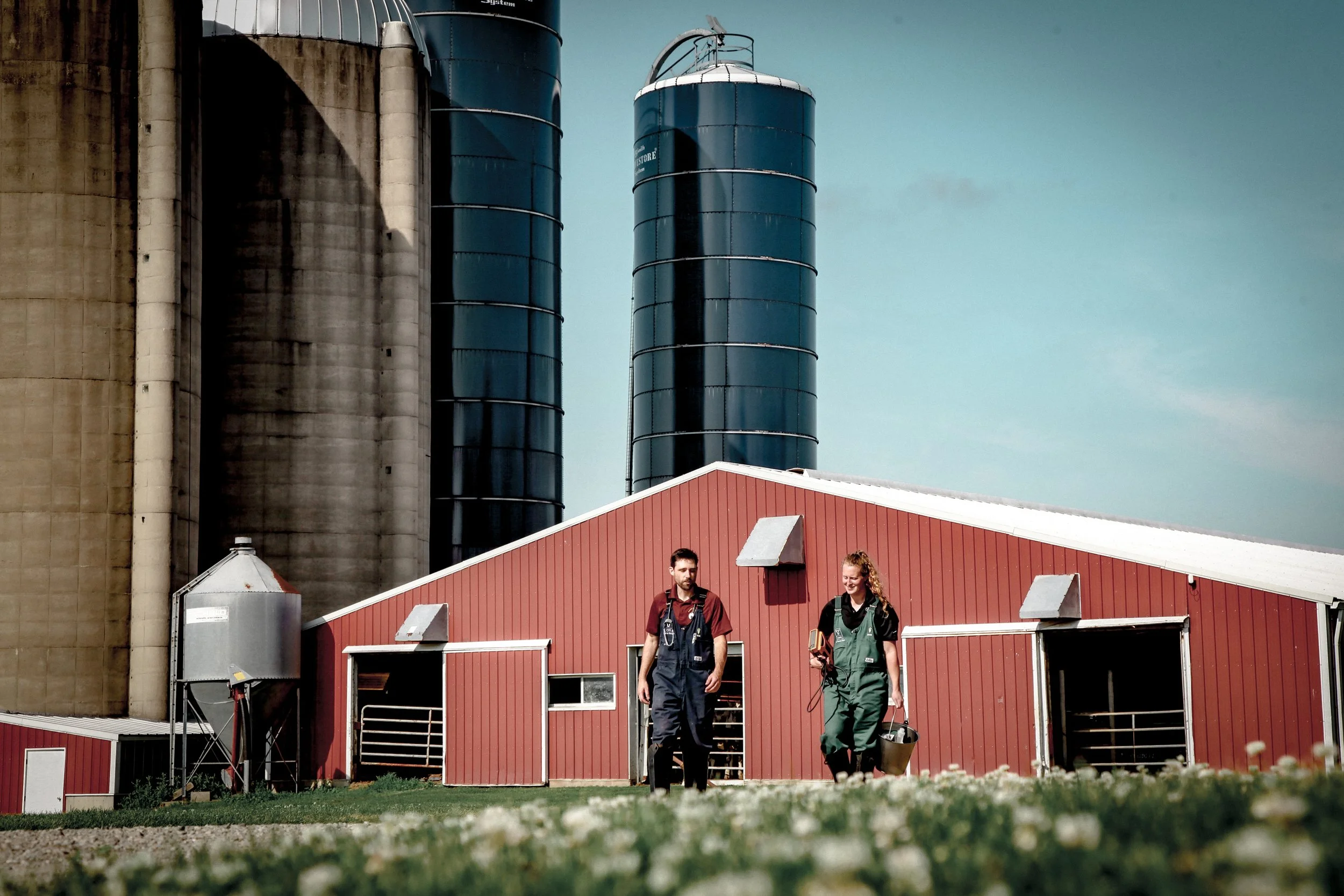 Two farmers walking outside a red barn on farmland with large silos in the background under a partly cloudy sky.
