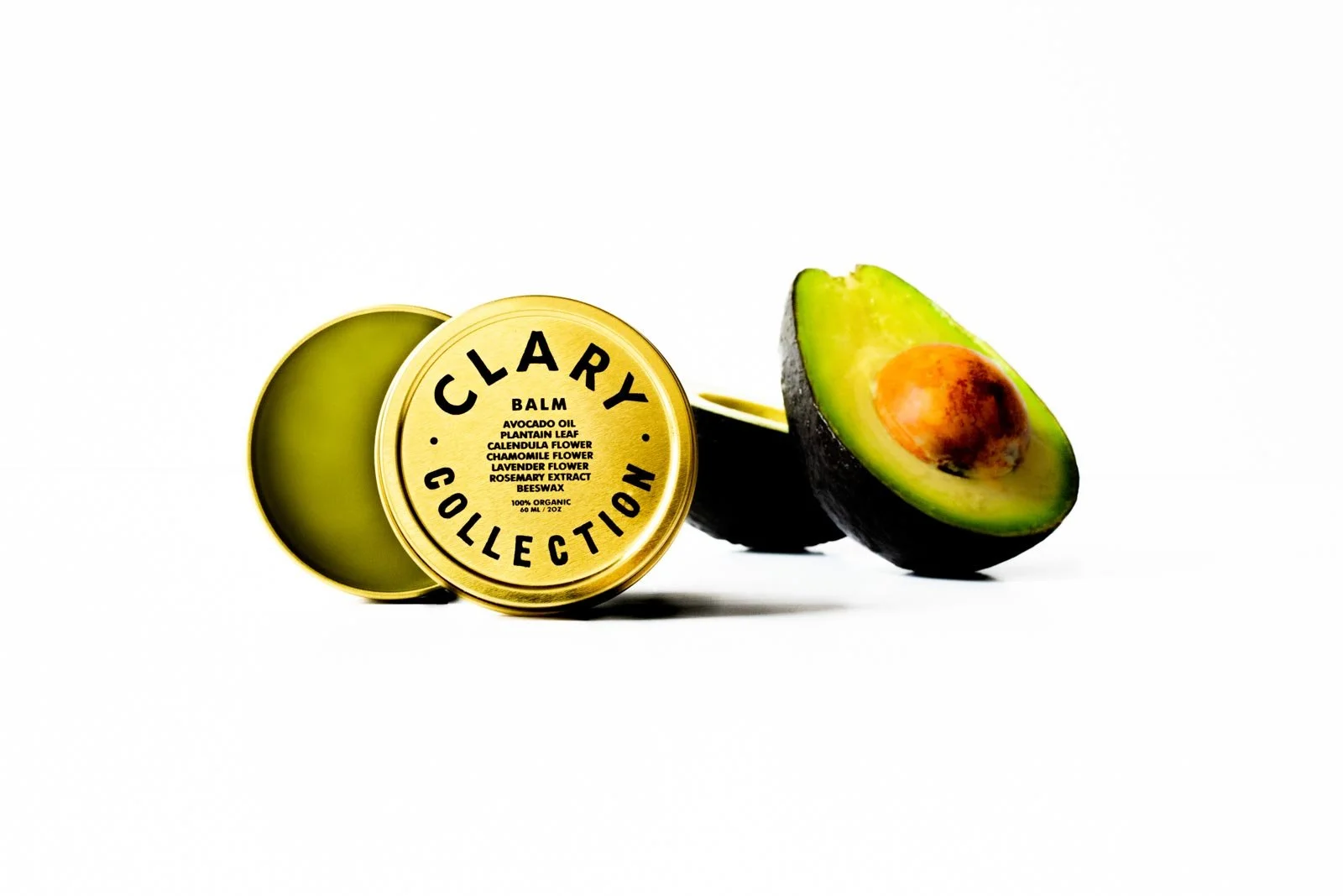 A small round tin labeled 'Clary Collection Balm' in front of a halved avocado with the seed visible on the right side of the image, set against a white background.
