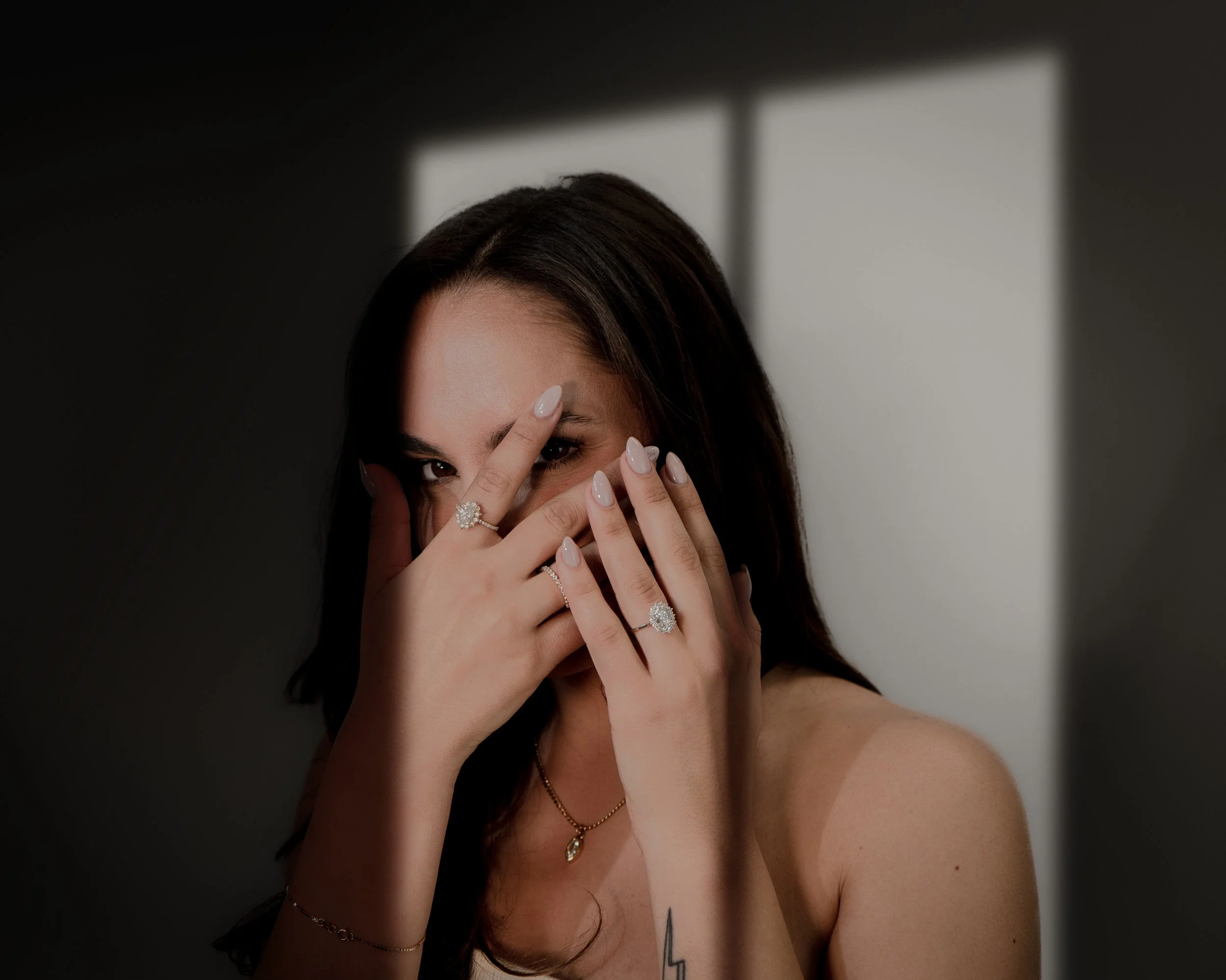 A woman with dark hair covering part of her face with her hands, wearing rings and a necklace, standing in front of a window casting a shadow.