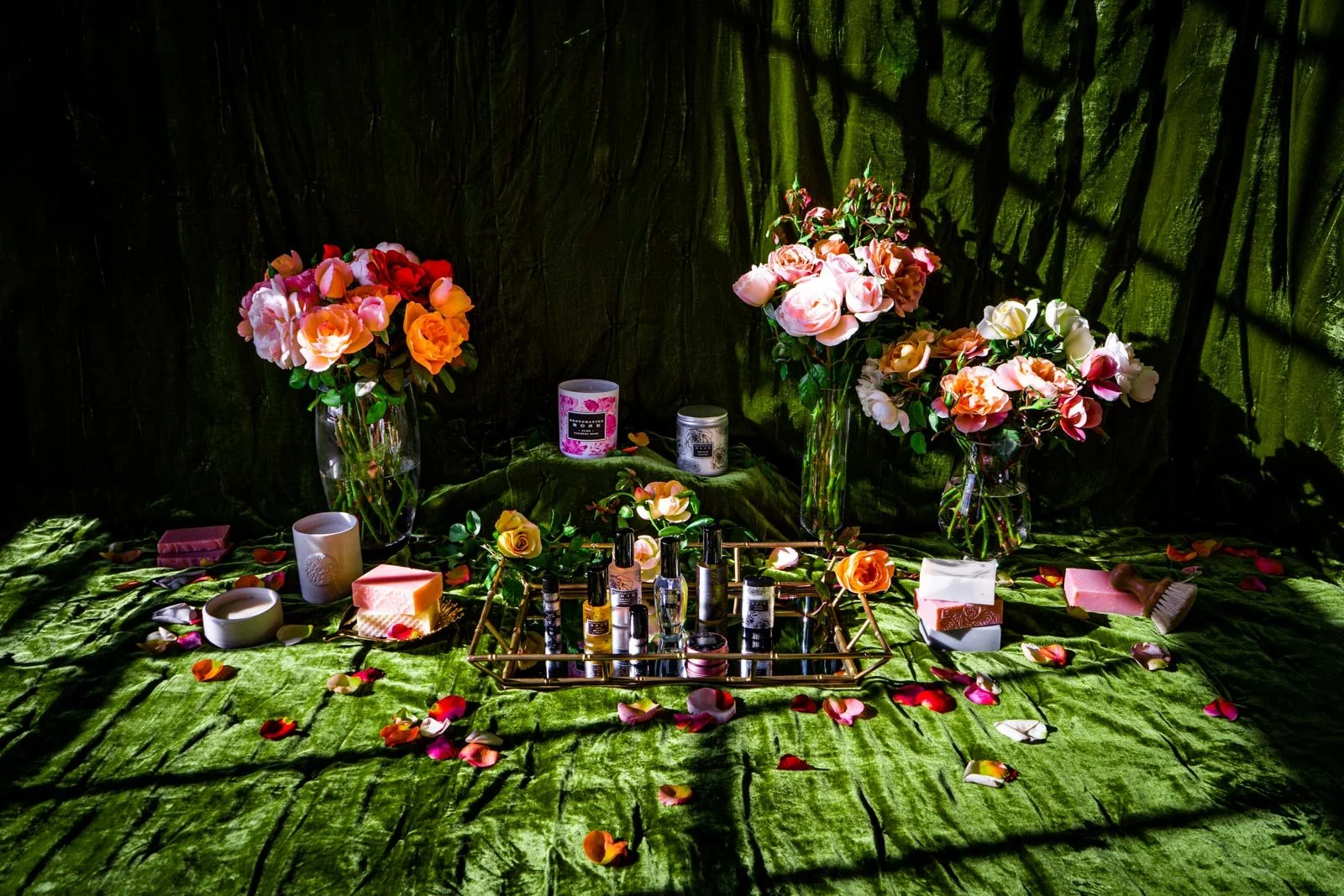 Arrangement of pink and peach roses in glass vases, along with nail polish bottles, candles, soap bars, and rose petals on a green textured surface with a dark background.