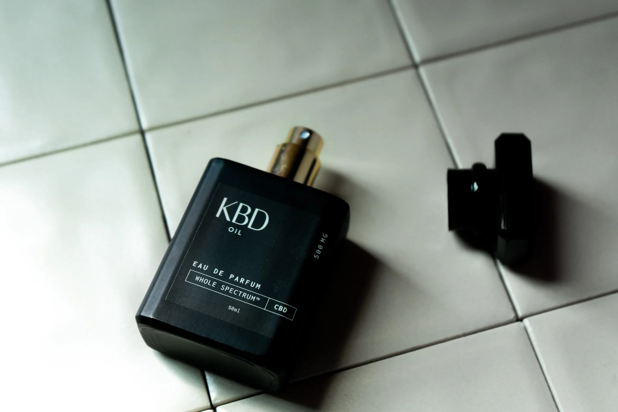 Bottle of CBD oil labeled 'KBD Oil' with a spray top, lying on a tiled floor next to a black object.