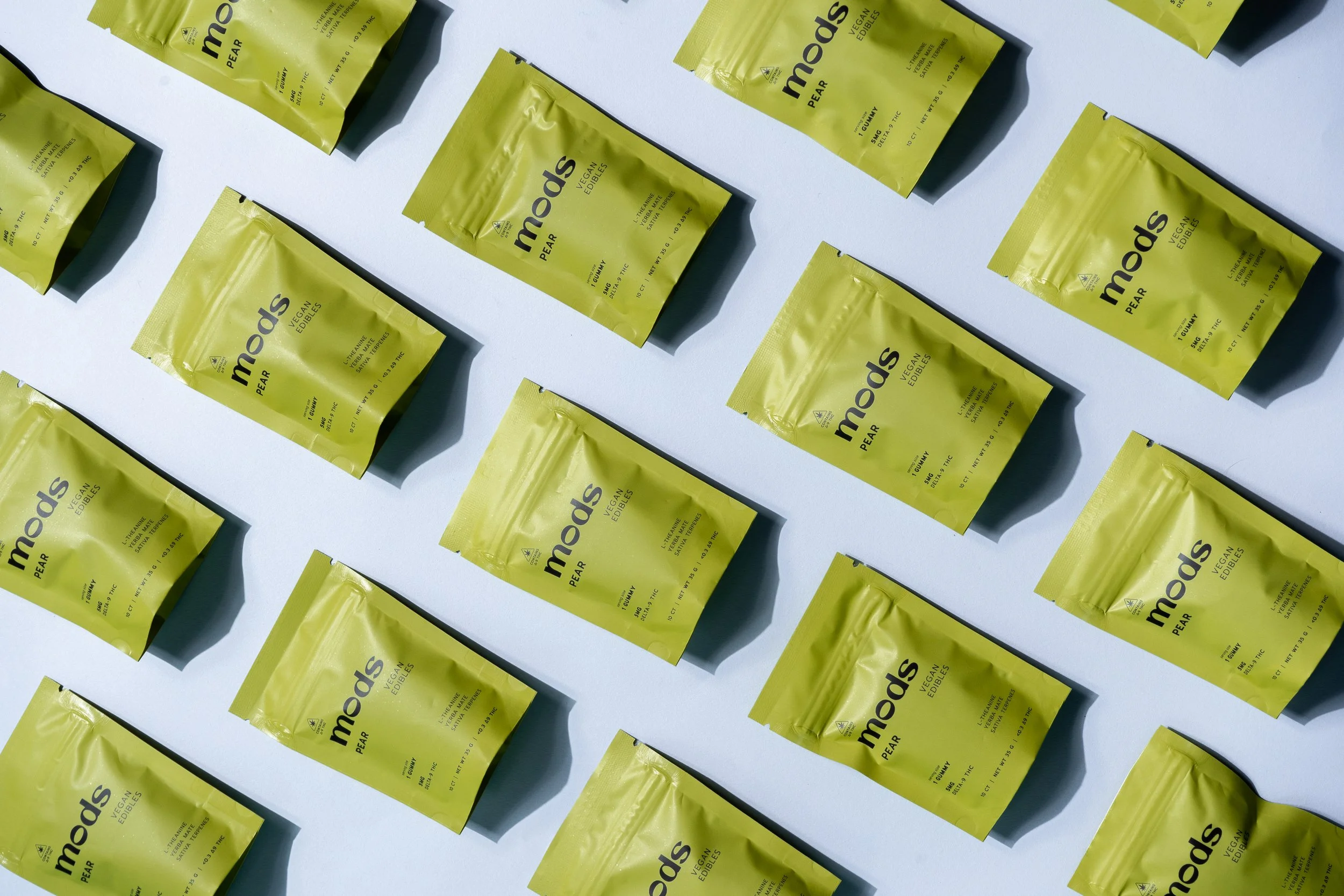 Multiple yellow snack bags labeled "mods PEAR VEGAN EDIBLES" arranged diagonally on a white background, casting shadows.