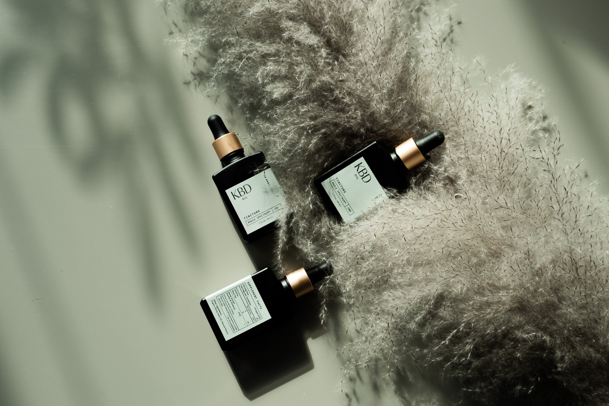 Three black bottles with white labels and gold caps placed on a light surface, surrounded by beige, fluffy dried plants.