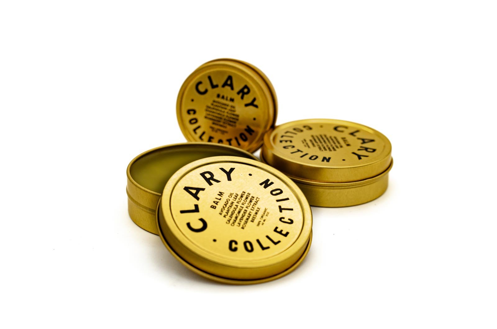 Open gold tin of Clarcy Collection balm with three closed tins beside it on a white background.